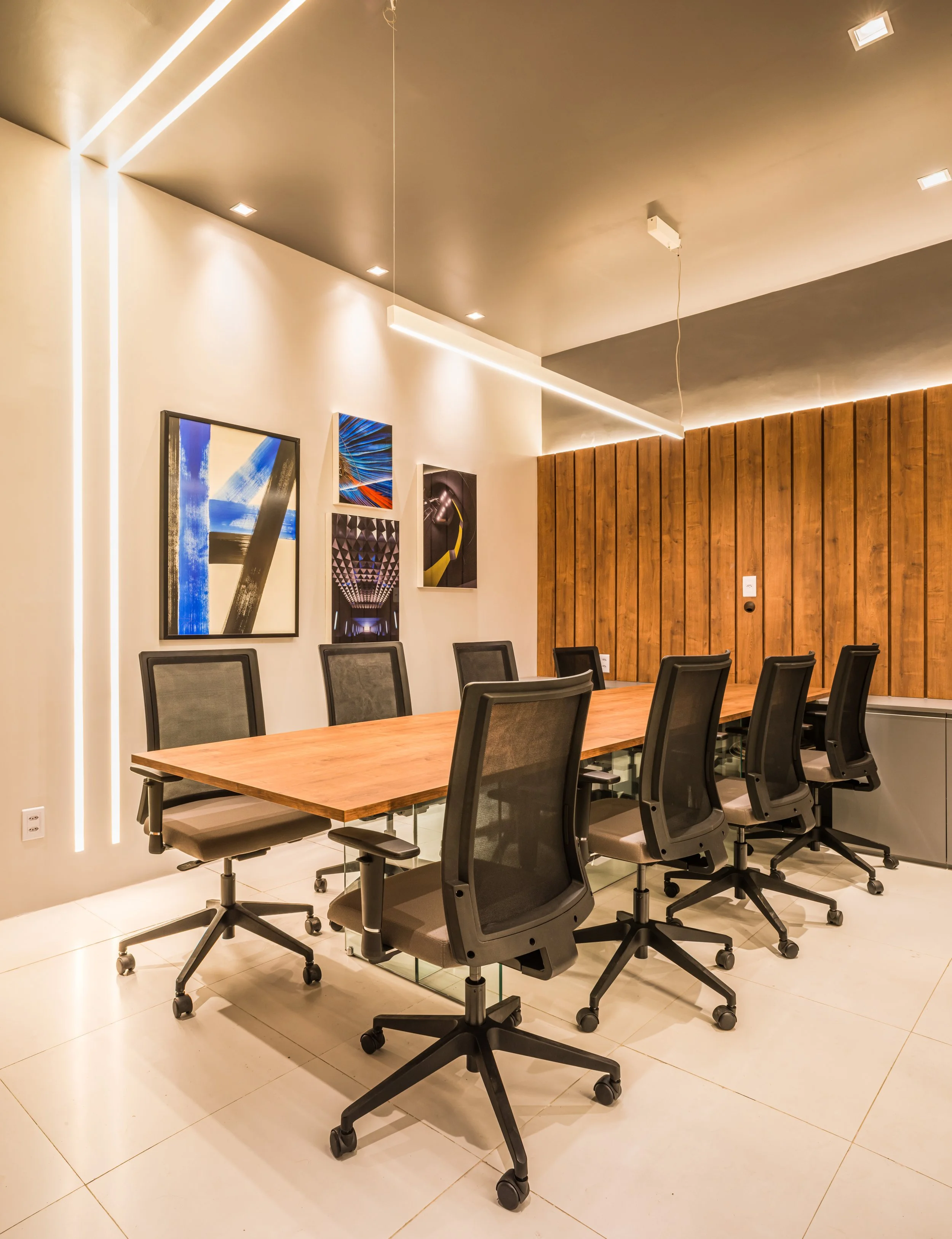 Modern conference room with a wooden table surrounded by eight black ergonomic office chairs, white tiled floor, white walls decorated with colorful abstract artwork, and a wooden accent wall with ceiling lights and LED strip lighting.