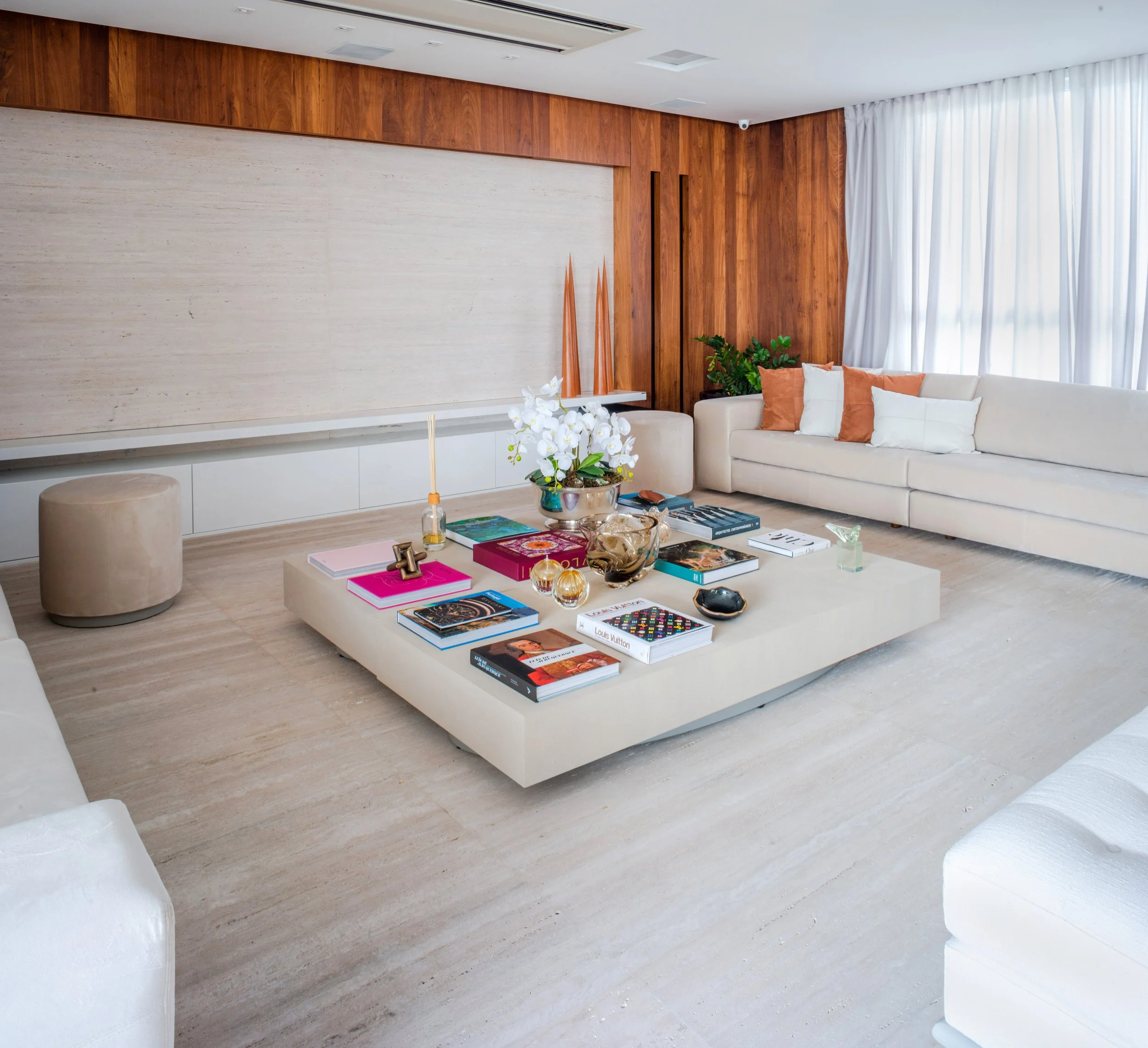 Modern living room with a large coffee table filled with books, decorative items, and flower arrangements, surrounded by white sofas and cream-colored ottoman.