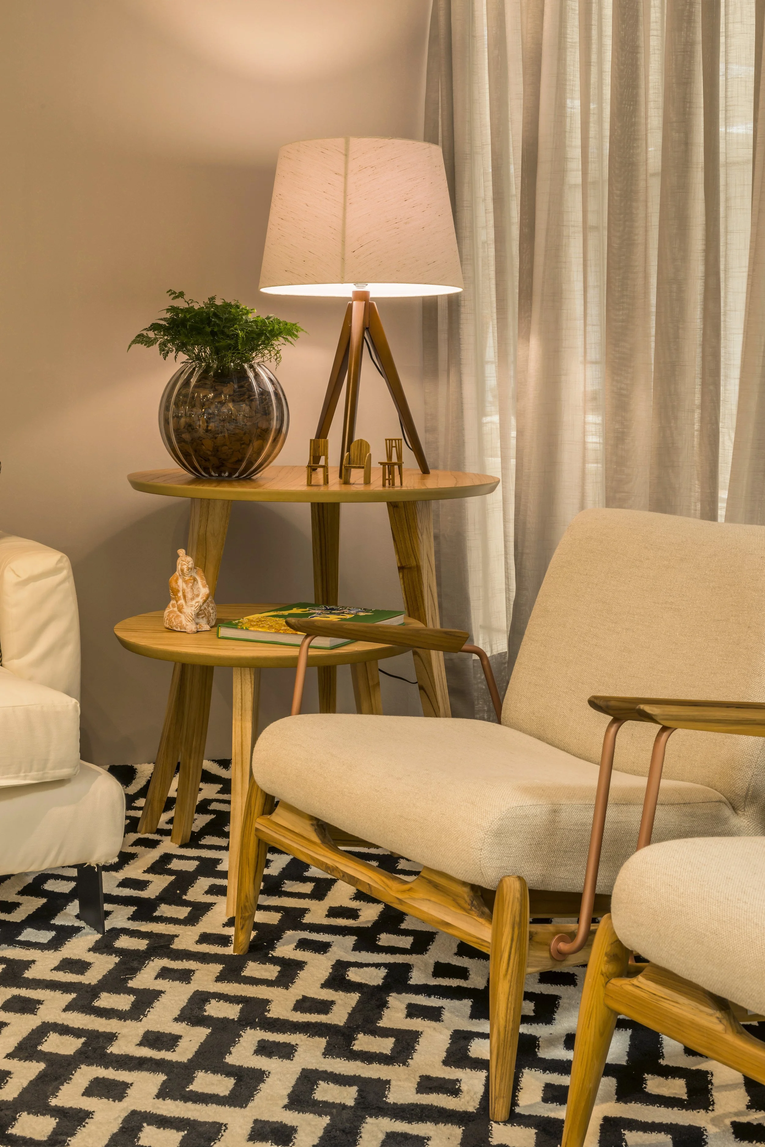Living room corner with a beige armchair, a black and white geometric-patterned rug, two wooden side tables with a lamp, a potted plant, a small figurine, and books.