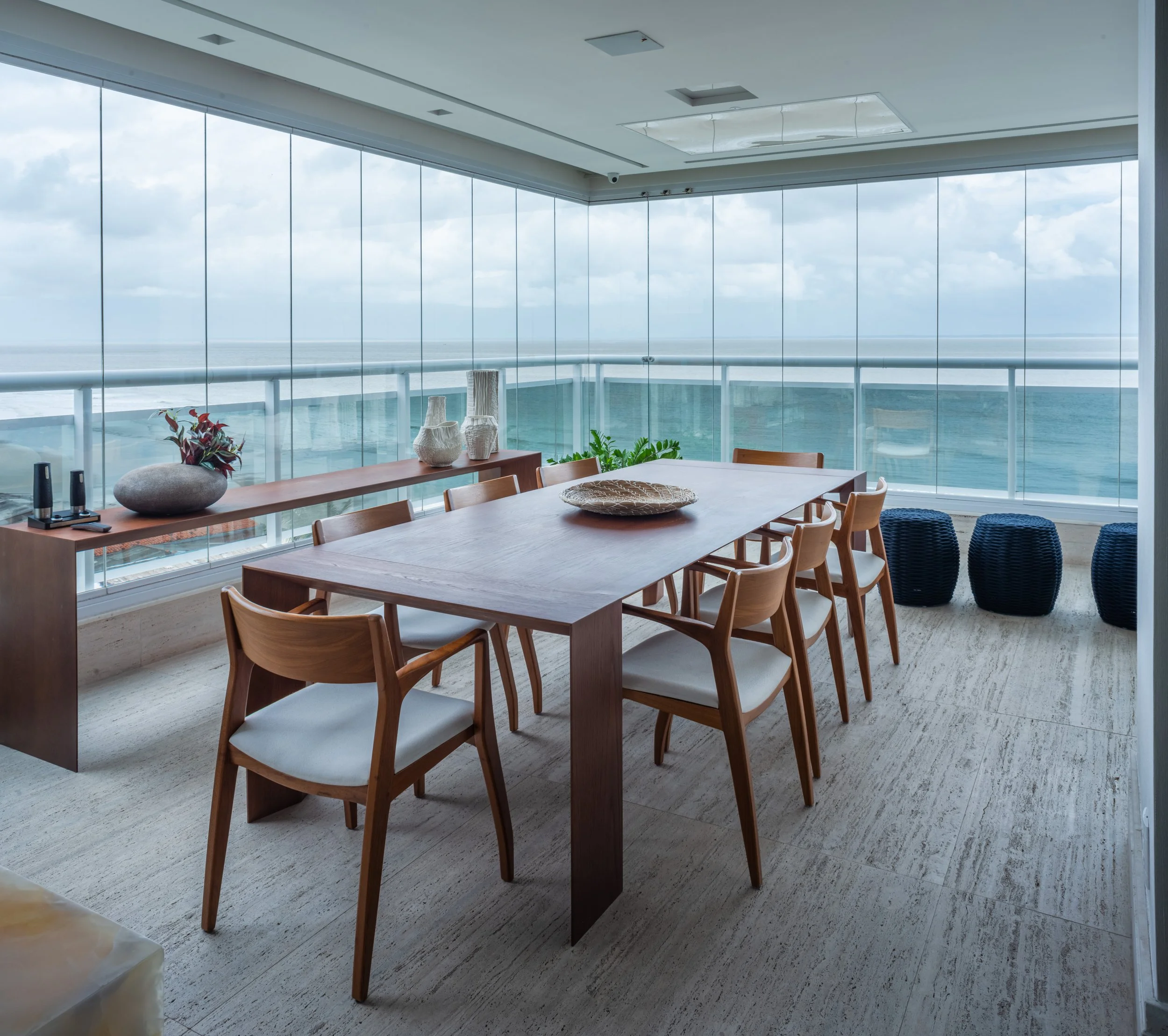 Empty dining room with large windows overlooking the ocean, wooden dining table with eight chairs, decorative items on a sideboard, and three black ottomans.