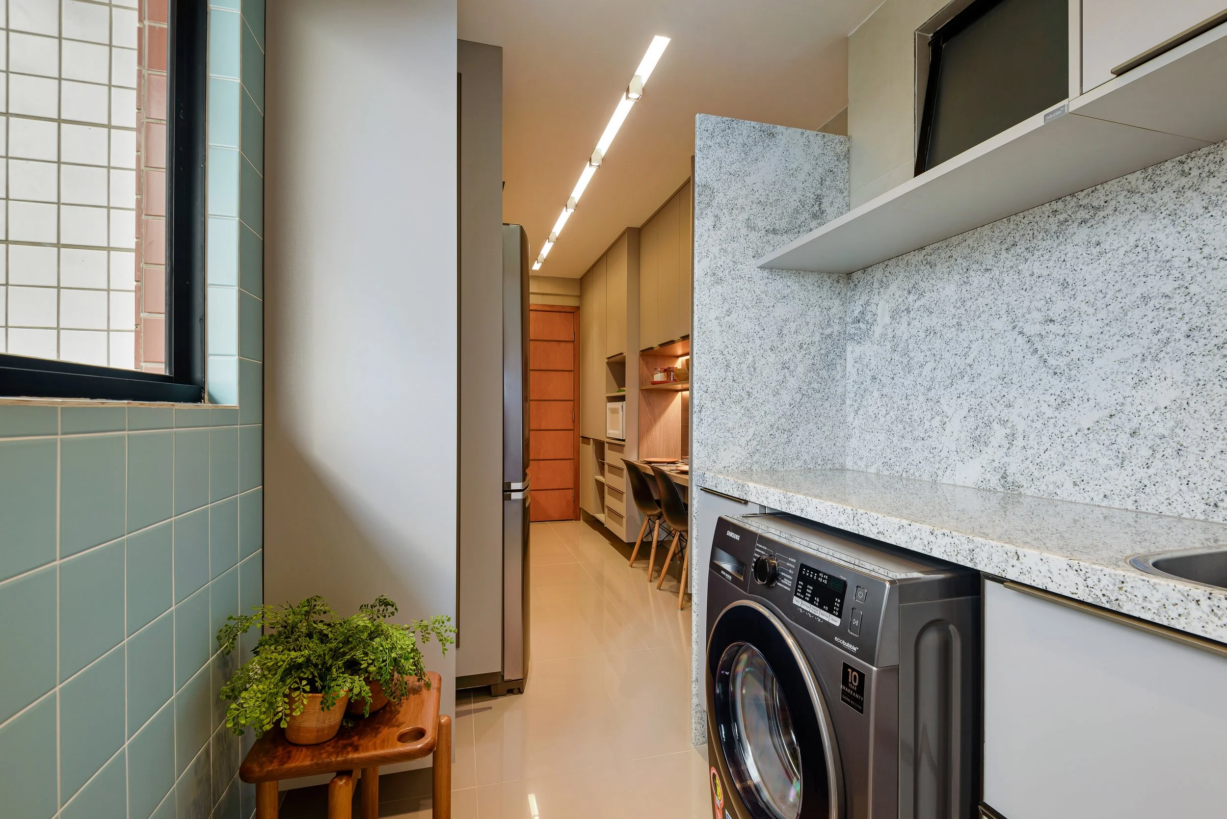 Modern kitchen with a washing machine, granite countertops, wooden cabinets, green tiled wall, and a small wooden table with a potted plant.