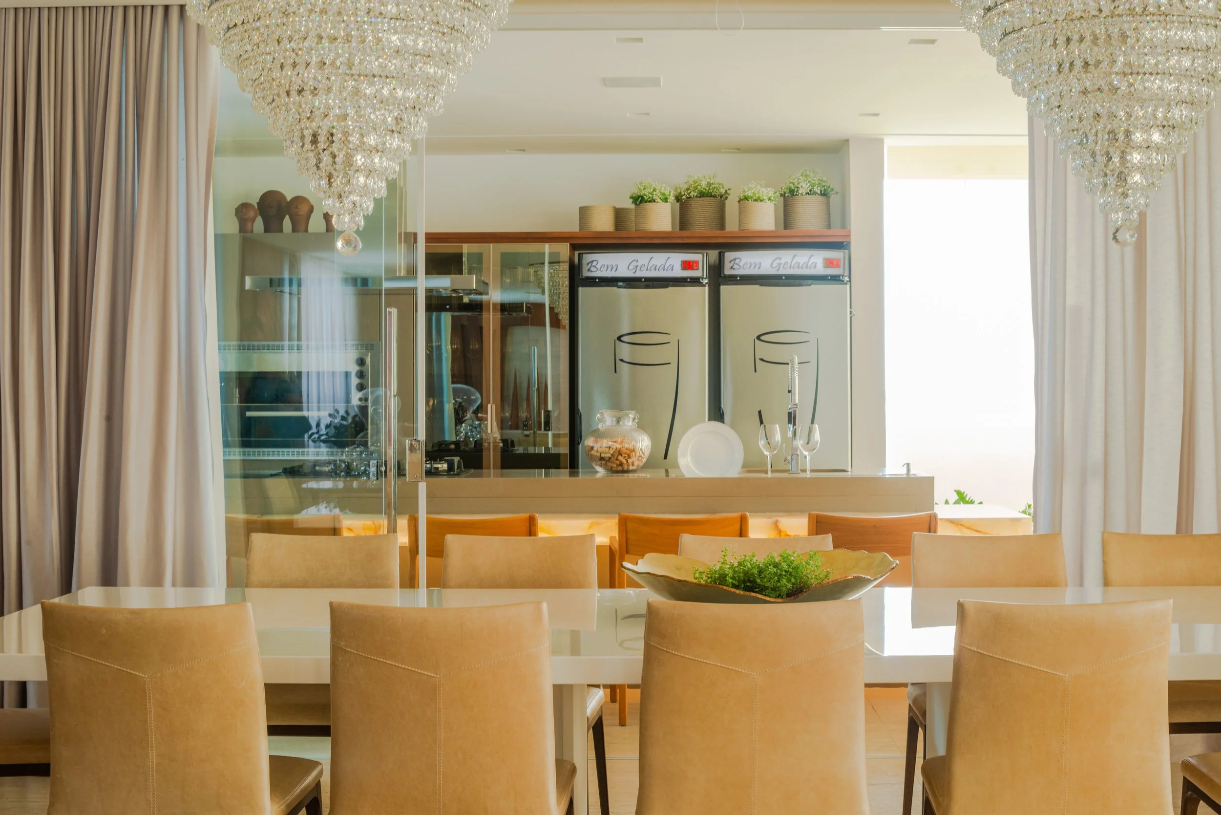 Elegant dining area with a large white table surrounded by beige chairs, a decorative green plant in a bowl, and a view of a kitchen with ice cream freezers, hanging chandeliers, and potted plants on a shelf.