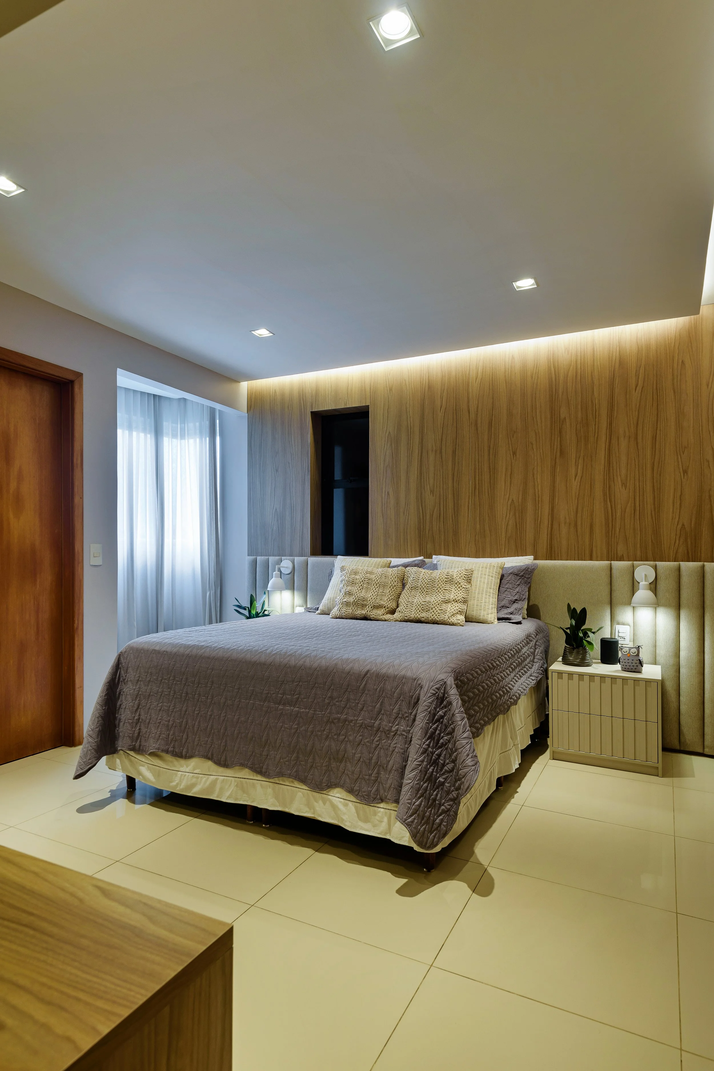 Modern bedroom with a large bed, beige headboard, wooden wall panel, side table with plants and lamps, white curtains, tiled floor, ceiling lights.
