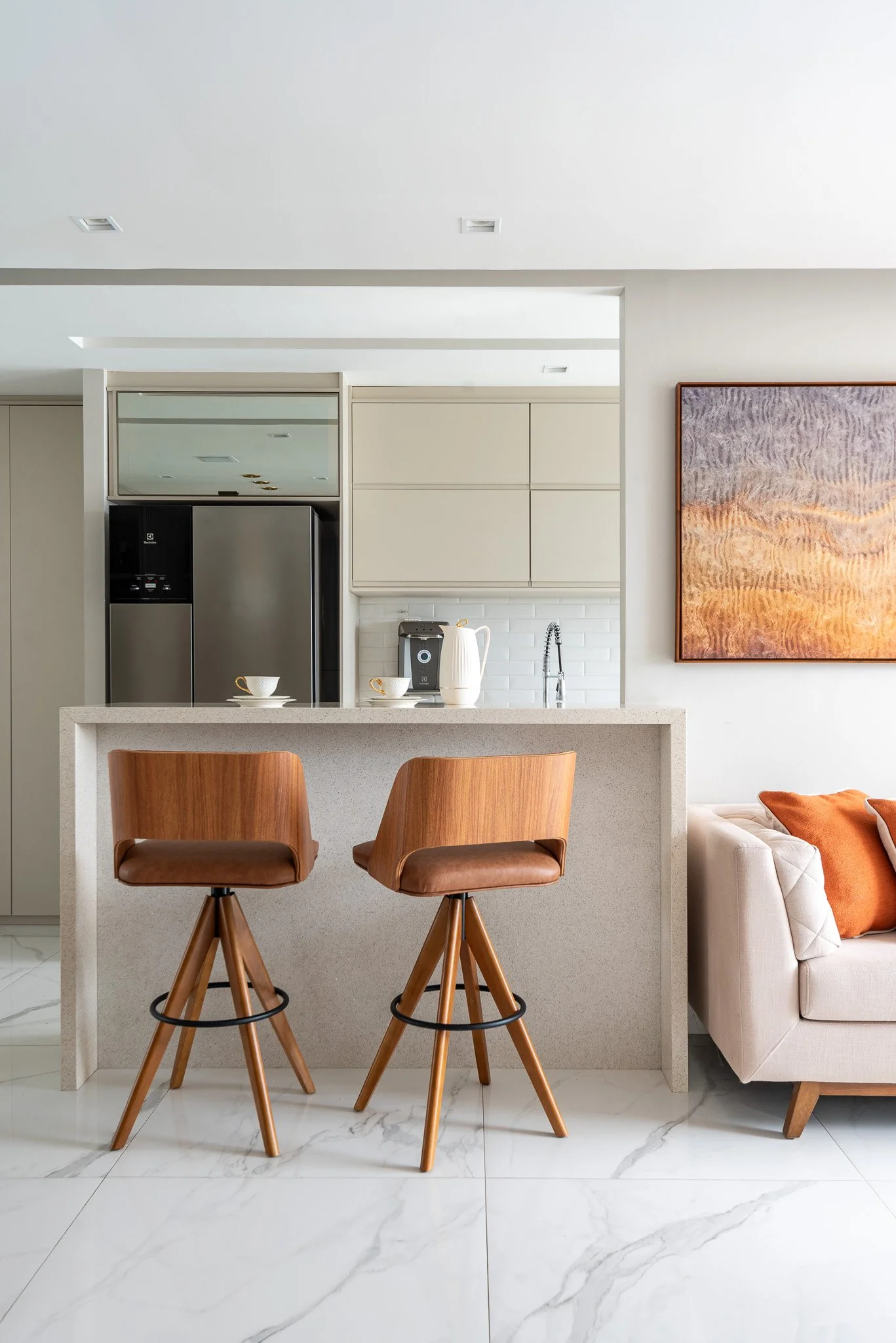 Modern kitchen with two wooden bar stools, beige countertop, white tile backsplash, stainless steel refrigerator, coffee maker, white teapot, and cups, adjacent to a living room with a white sofa and orange pillows, and a large abstract painting on t