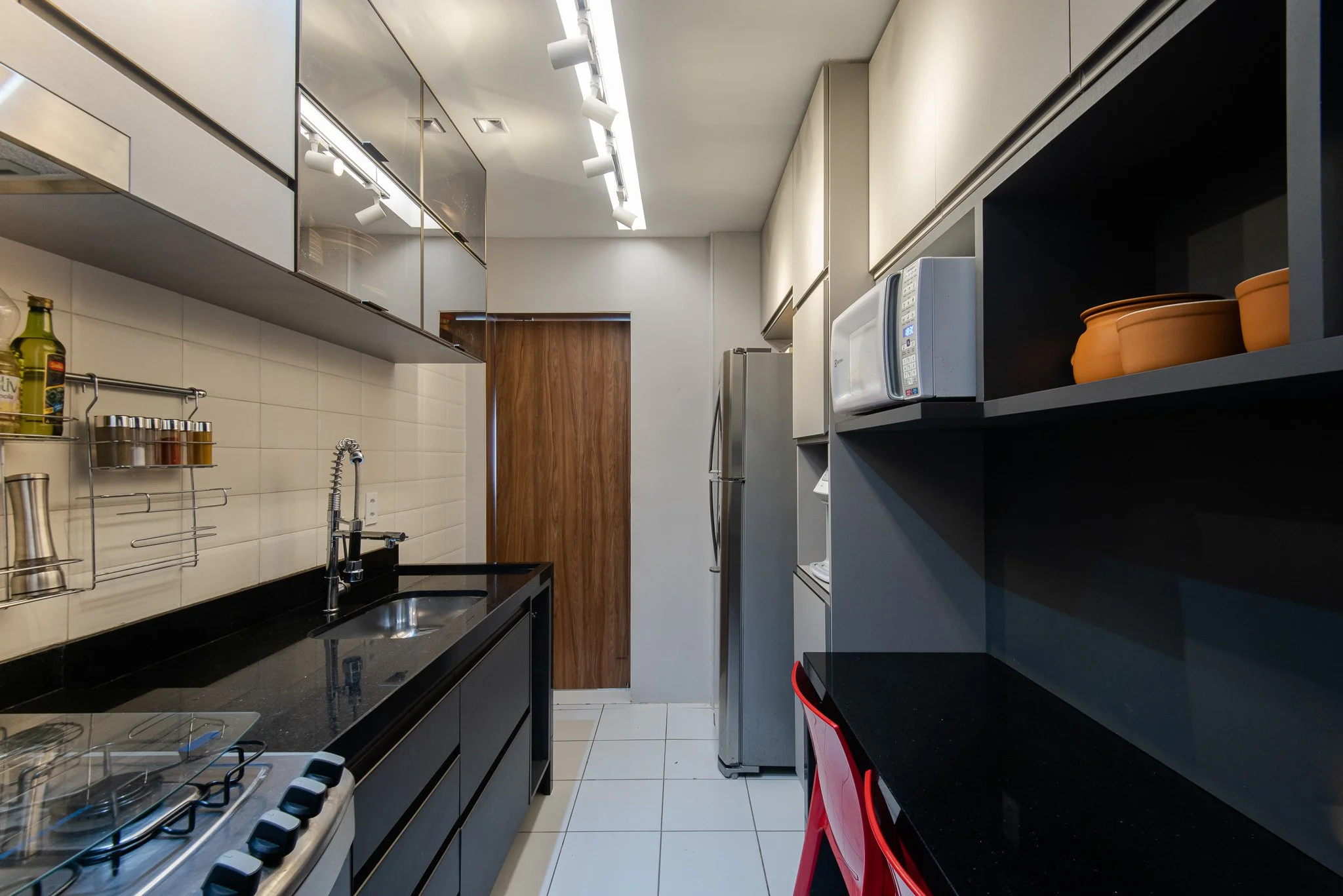 Modern narrow kitchen with black countertops, white cabinets, a stainless steel refrigerator, microwave, open shelves with terracotta pots, and a small red chair.