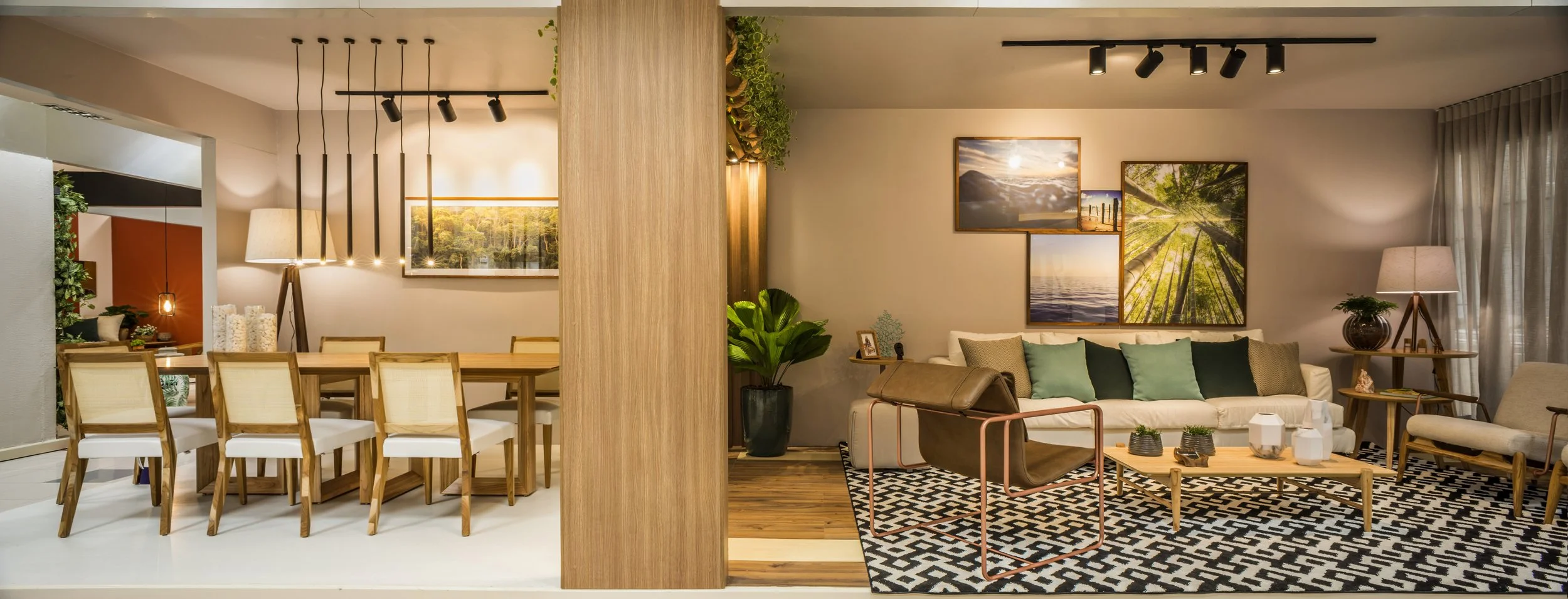 A living room and dining area separated by a wooden column in a modern apartment. The dining space features a large wooden table with eight cream-colored chairs, hanging pendant lights, and wall art. The living area has a white sofa with green cushio