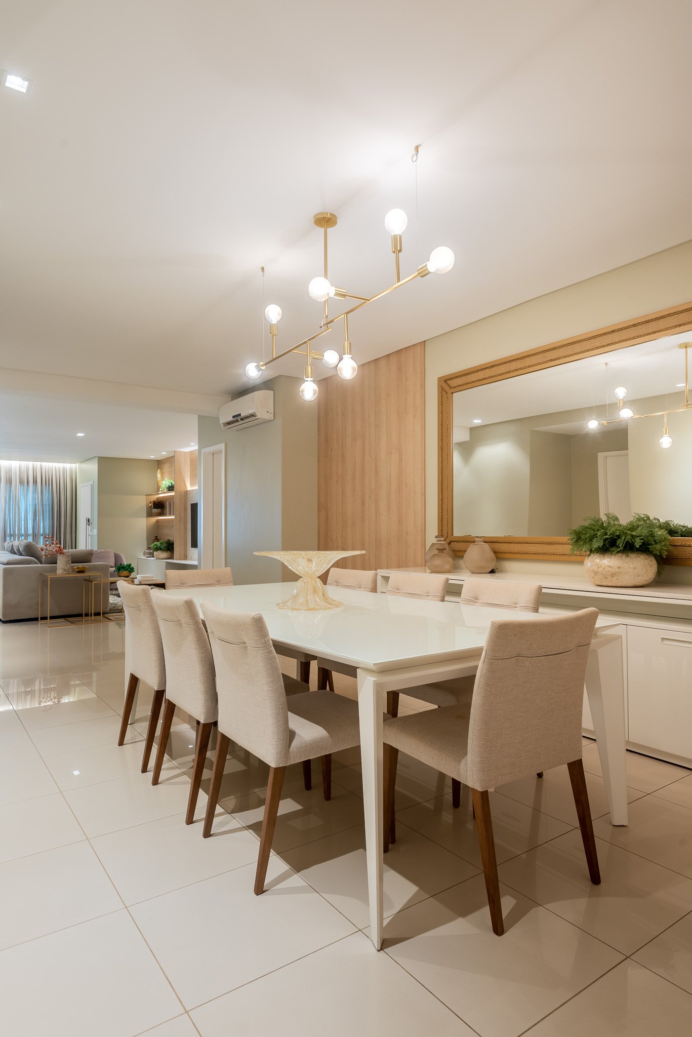 Modern dining room with a white table, beige upholstered chairs, large mirror, pendant lights, and decorative vases holding plants.