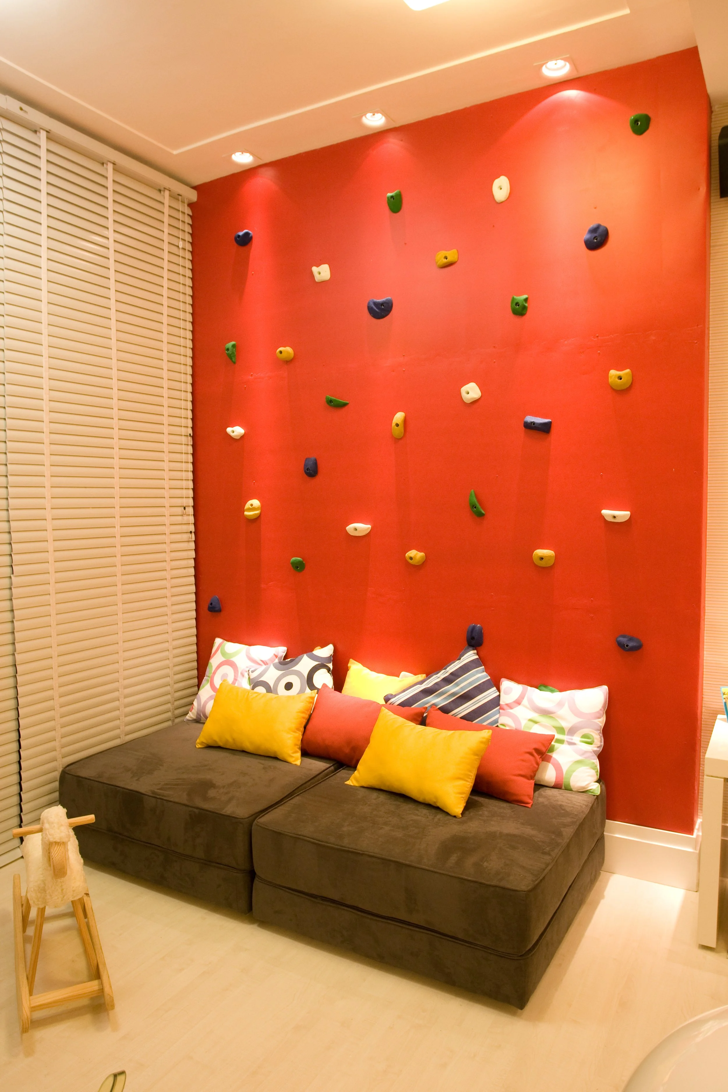 A red indoor climbing wall with colorful holds, positioned above a dark brown sofa with multiple yellow, red, and patterned pillows in a room with wooden flooring and white wall blinds.