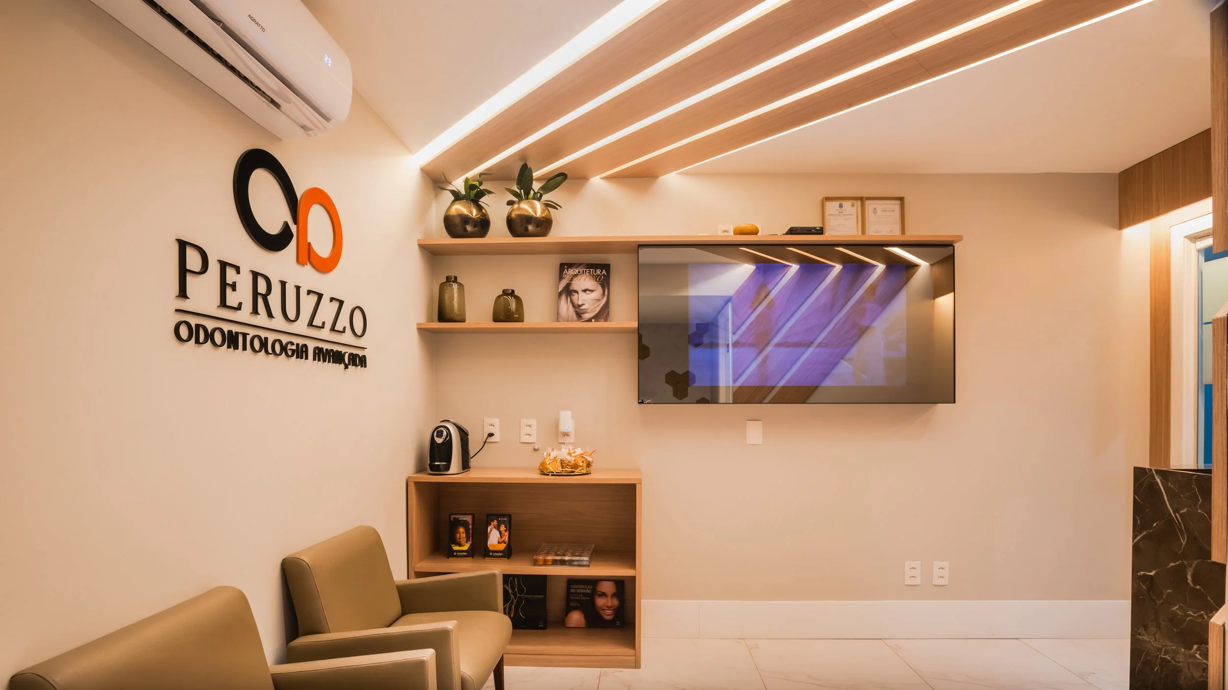 Interior of a modern dental office with a beige wall, decorative shelves with vases, a mounted flat-screen TV, and waiting chairs.