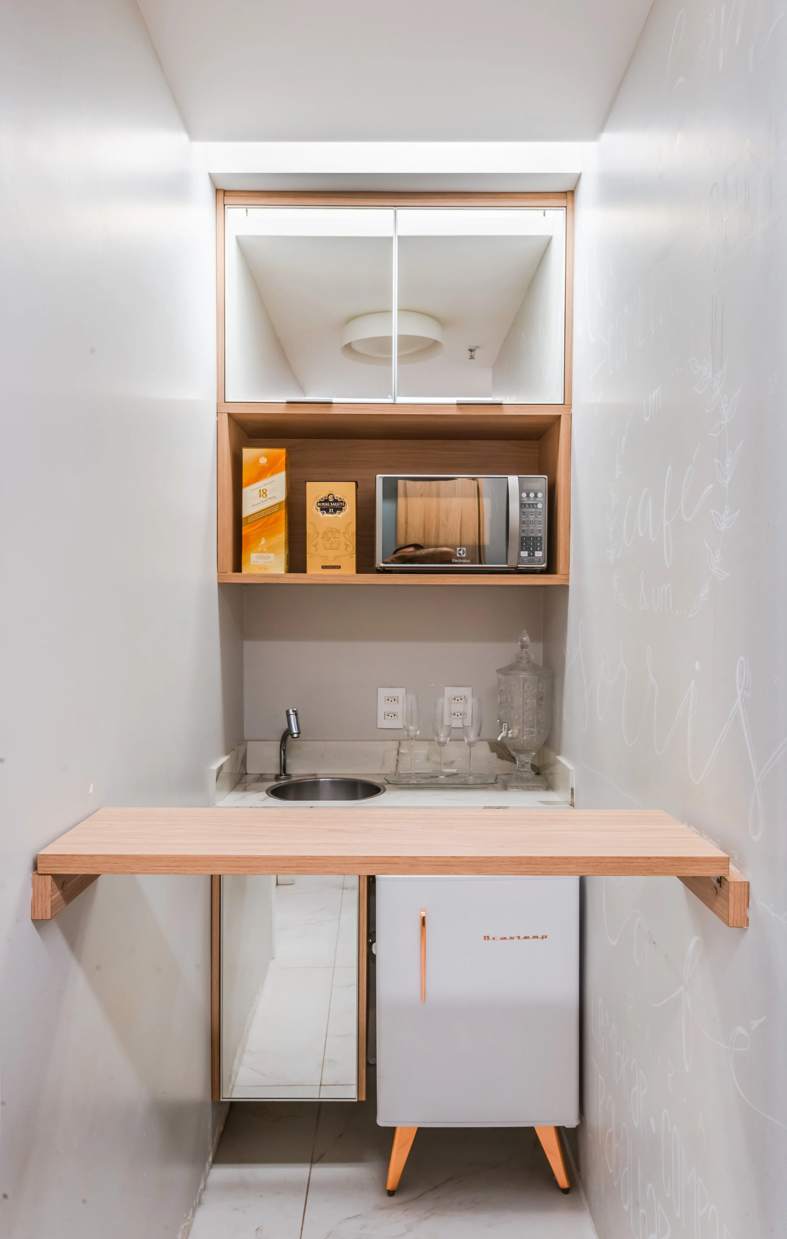 Small kitchenette with a mini refrigerator, microwave, and shelf with boxes on it. The kitchenette has a marble countertop, a small sink, and is framed in light wood. There are wall outlets above the countertop, and a mirror partially visible on the 
