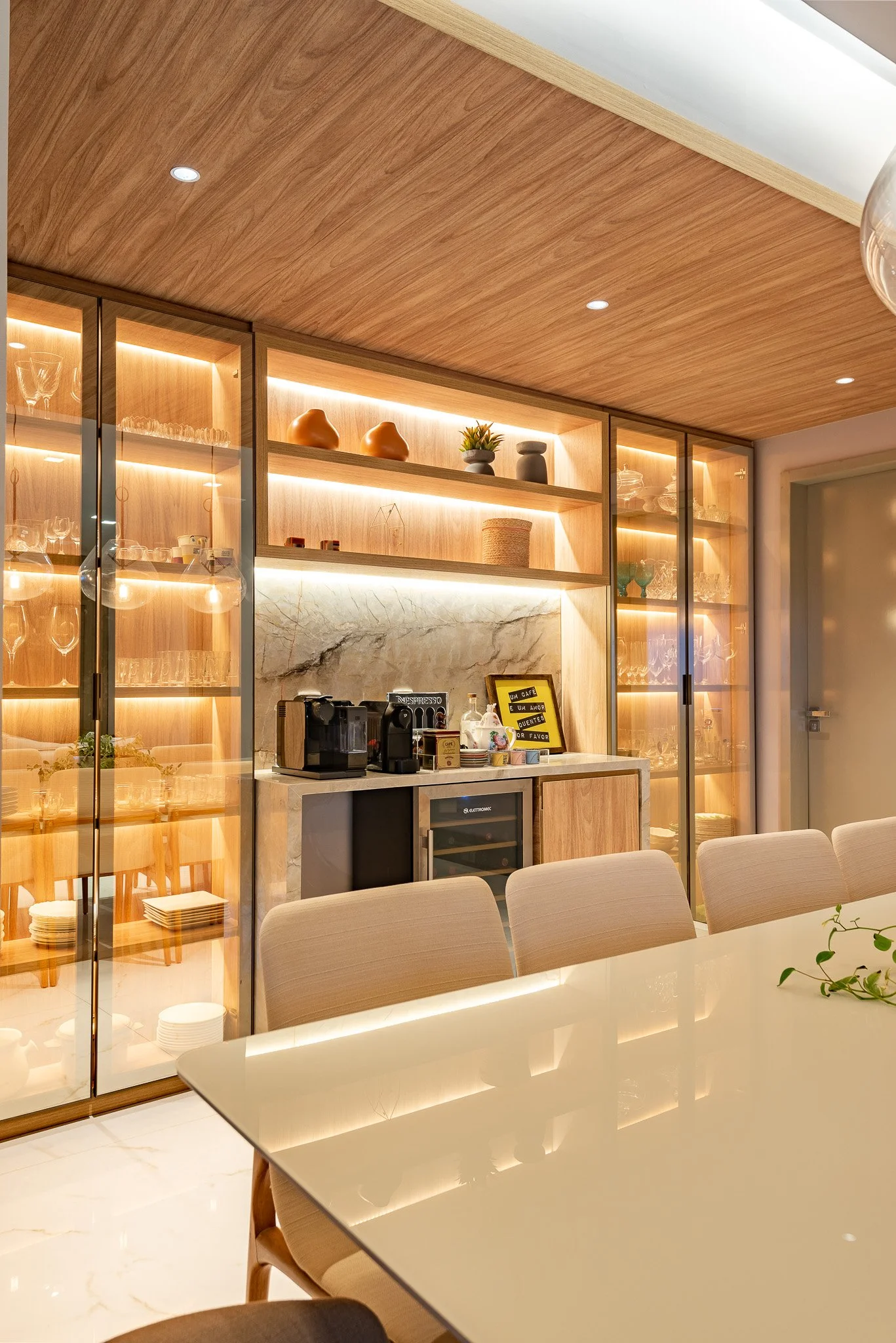 A modern dining area with a white table, beige chairs, and a wooden wall with glass cabinet doors filled with glassware. There is a coffee station with a coffee machine, mini fridge, and decorative items on shelves. The ceiling is wooden with recesse
