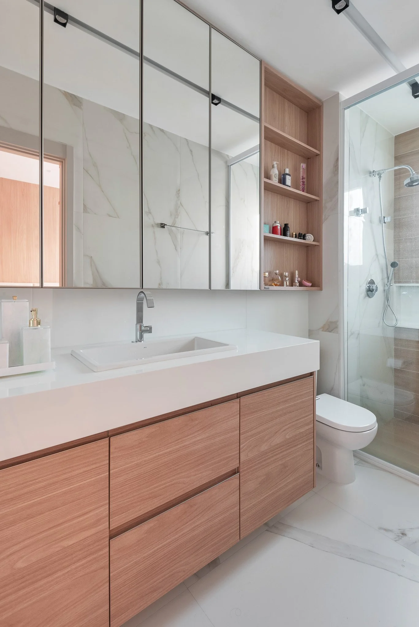 Modern bathroom with wood cabinet vanity, white countertop, large mirror cabinets, glass shower with brown accent tiles, and wooden open shelves with toiletries.