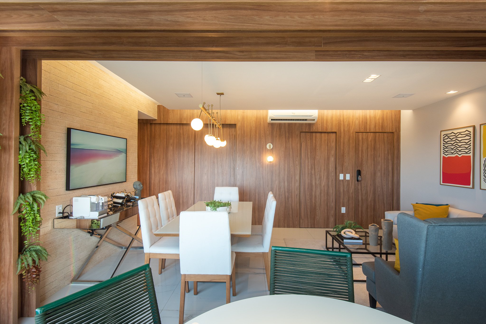 Modern living room with wooden walls, white chairs around a dining table, a blue armchair, hanging pendant lights, framed artwork on the wall, and green plants.