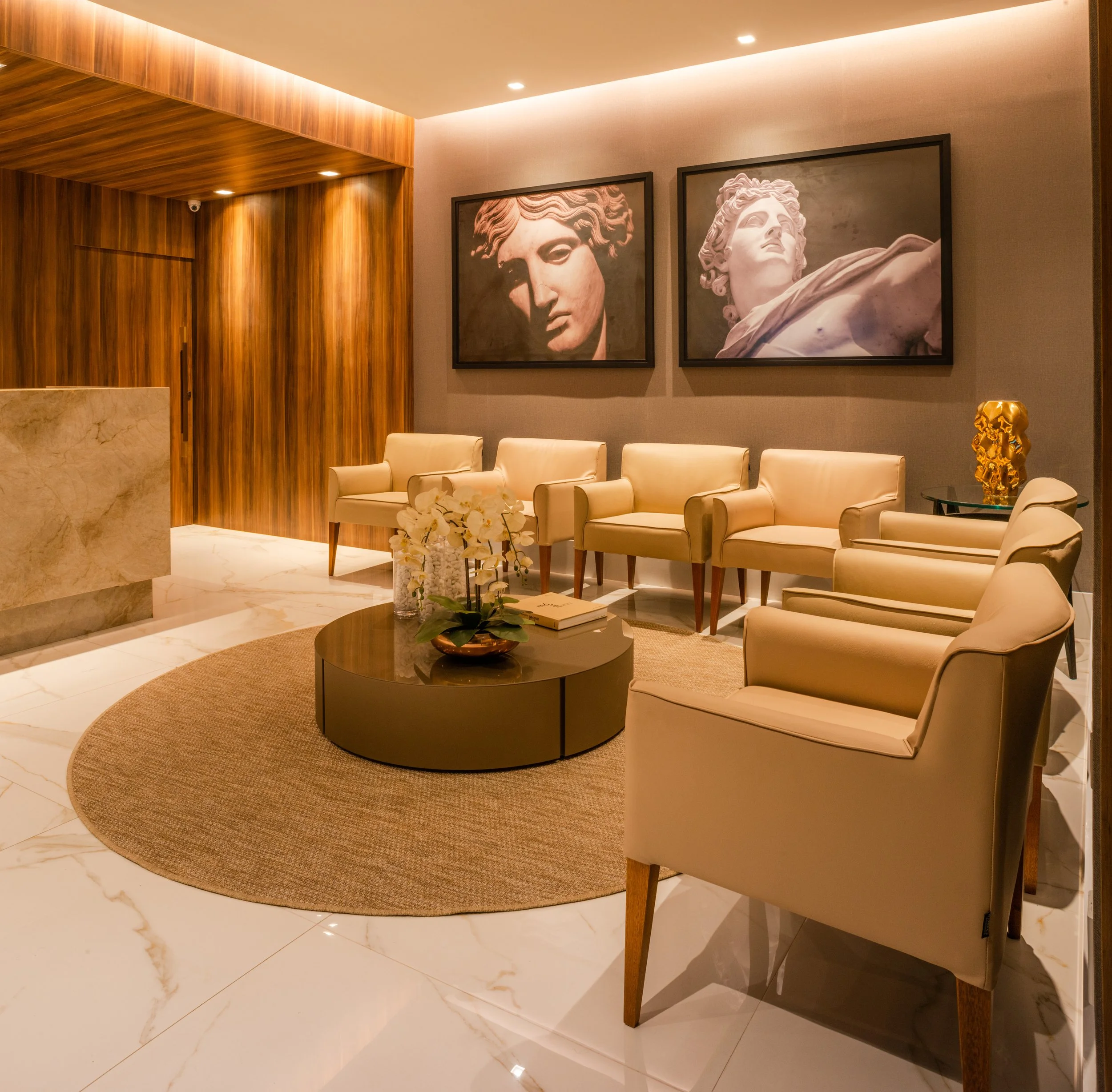 A modern hotel lobby sitting area with cream-colored armchairs and a round coffee table decorated with flowers and books. Two large black-and-white framed artwork of classical sculptures hang on the wall, and a gold decorative sculpture is on a side 