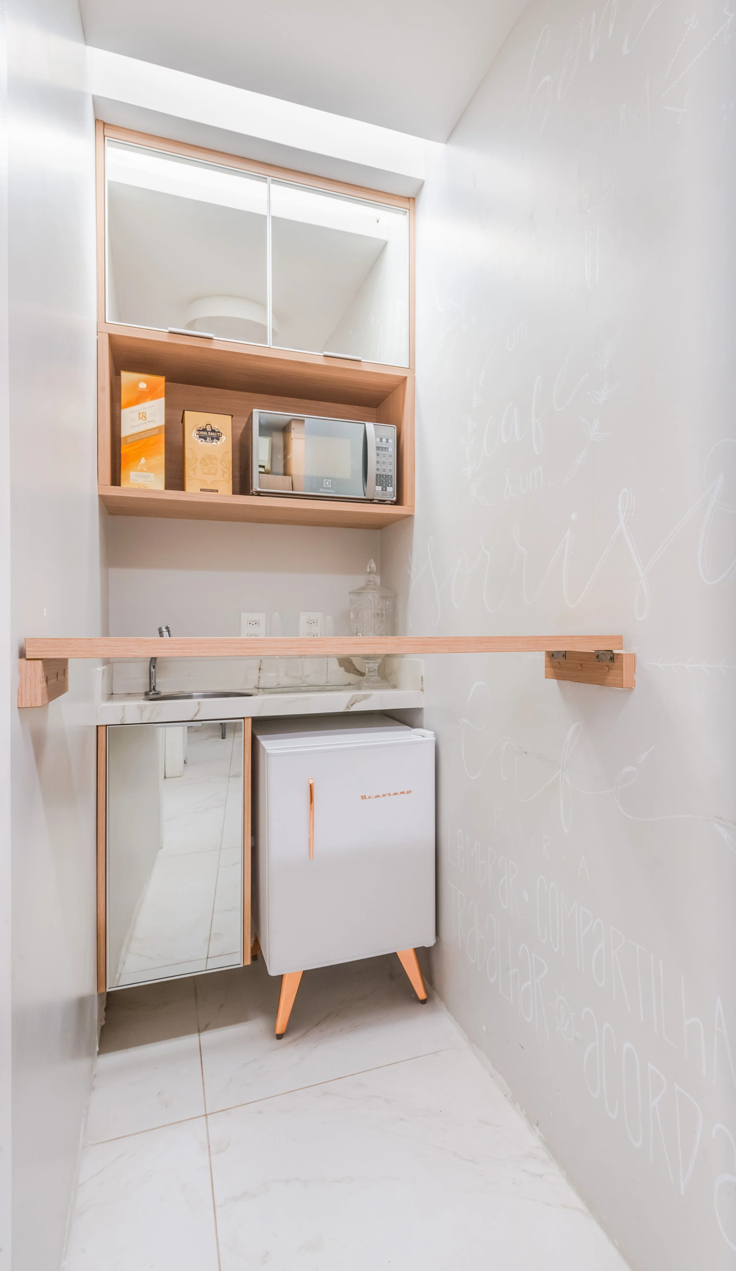 Small kitchen area with a microwave, mini fridge, countertop, and wall shelves, featuring a wall with white chalk handwriting.