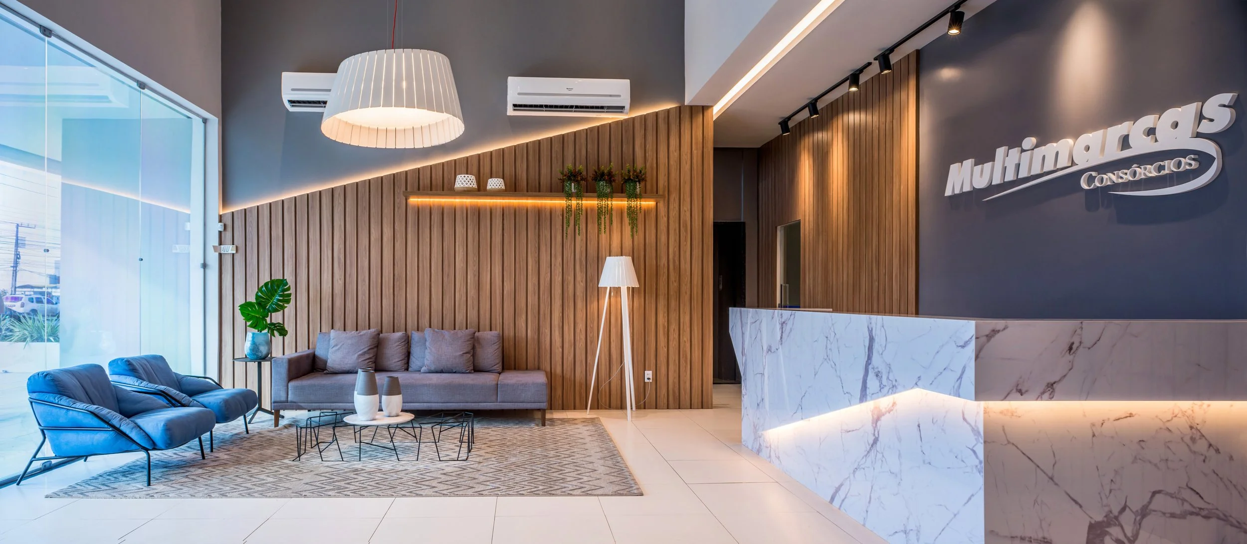 Modern hotel lobby with a front desk, wooden wall accent, gray sofa, blue lounge chairs, a patterned rug, and decorative plants and vases, with large window letting in natural light.