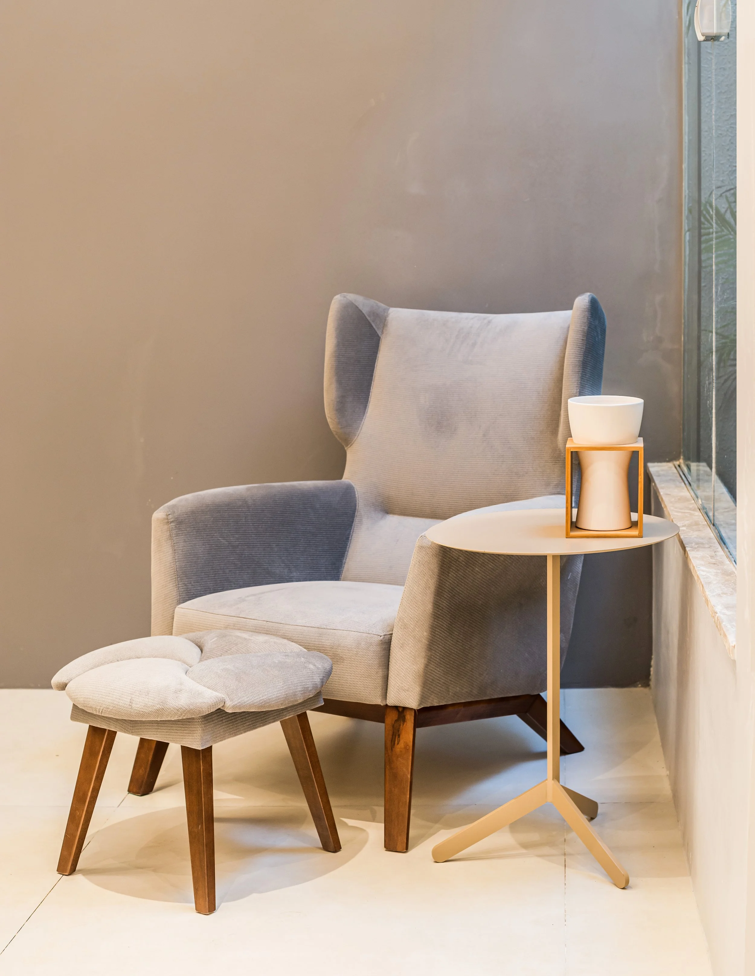 A cozy reading nook with a modern wingback chair in gray with wooden legs, a matching footstool, and a small beige side table with a ceramic bowl and a decorative frame, positioned beside a large window with urban view.