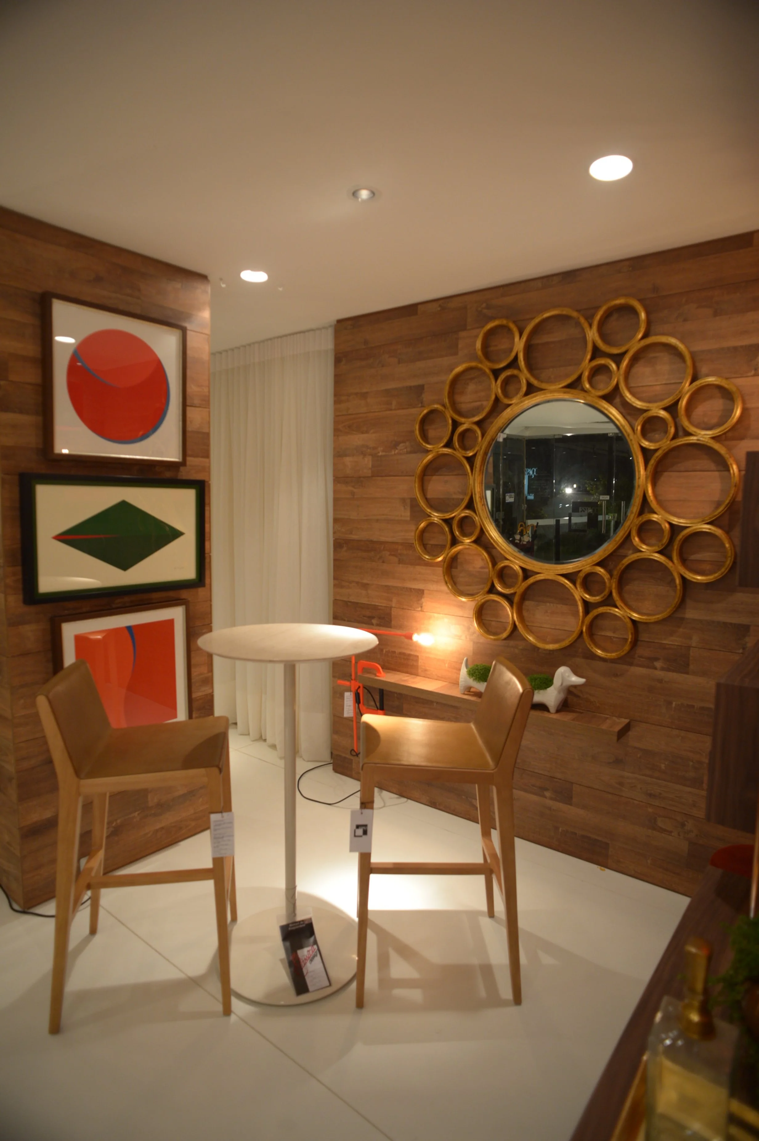 Interior of a modern, well-lit room with wooden walls, two wooden chairs around a small round table, framed abstract artwork on a wall, and a large decorative circular mirror.