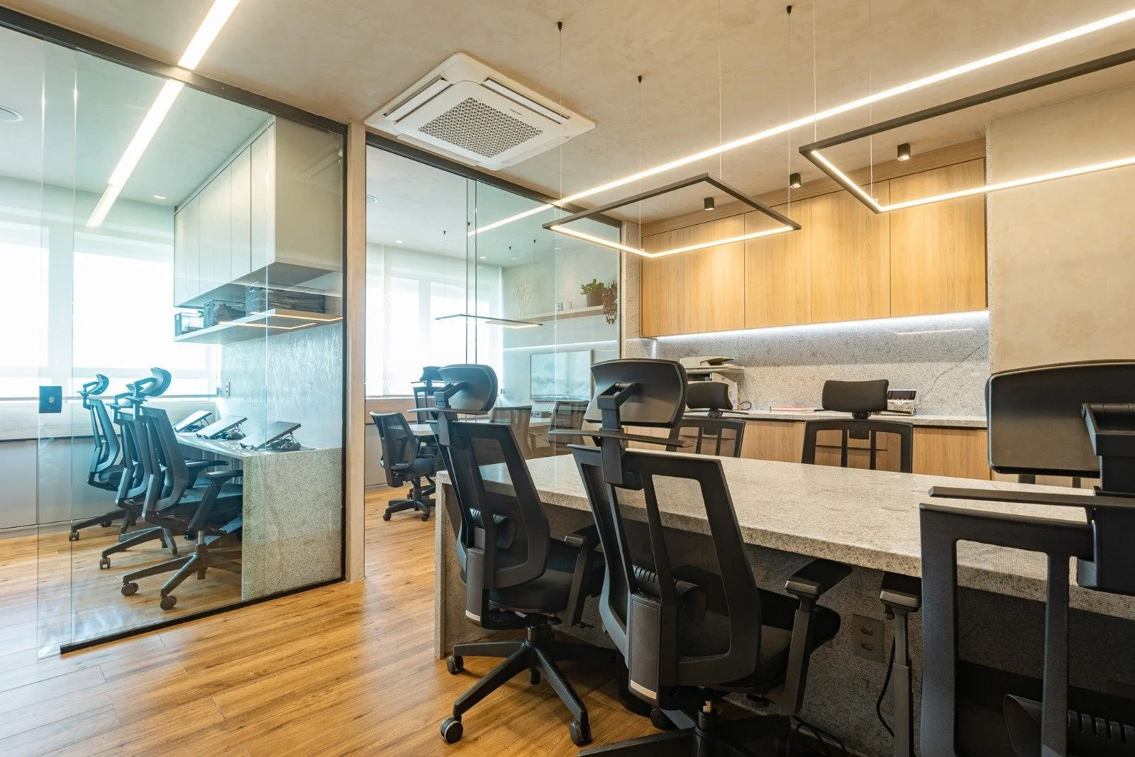 Modern office space with glass-walled conference room, black ergonomic office chairs, wood and granite countertops, overhead lighting, and a ceiling vent.