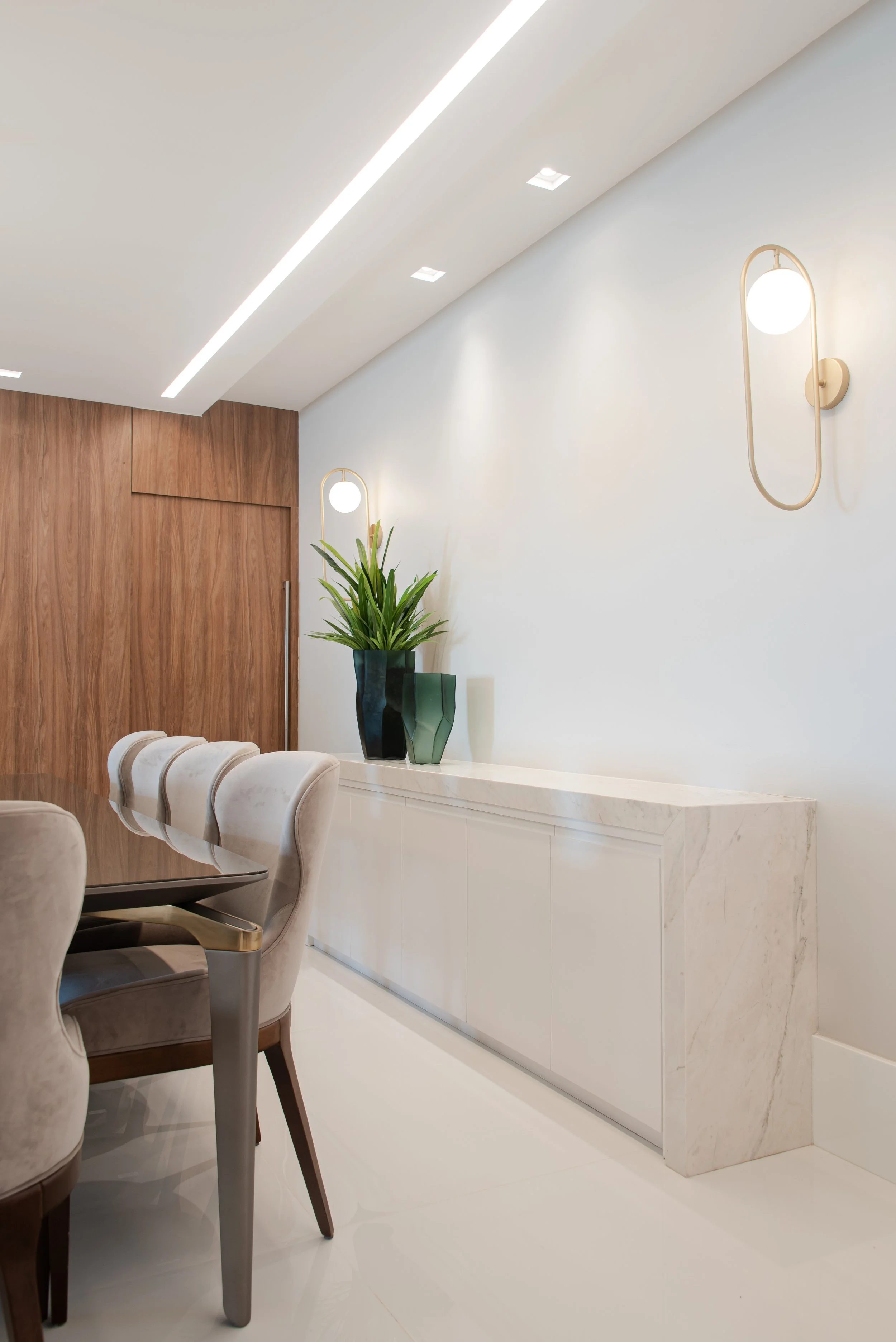 Modern interior space with a white marble sideboard, potted plants, upholstered chairs, and wall-mounted lamps.