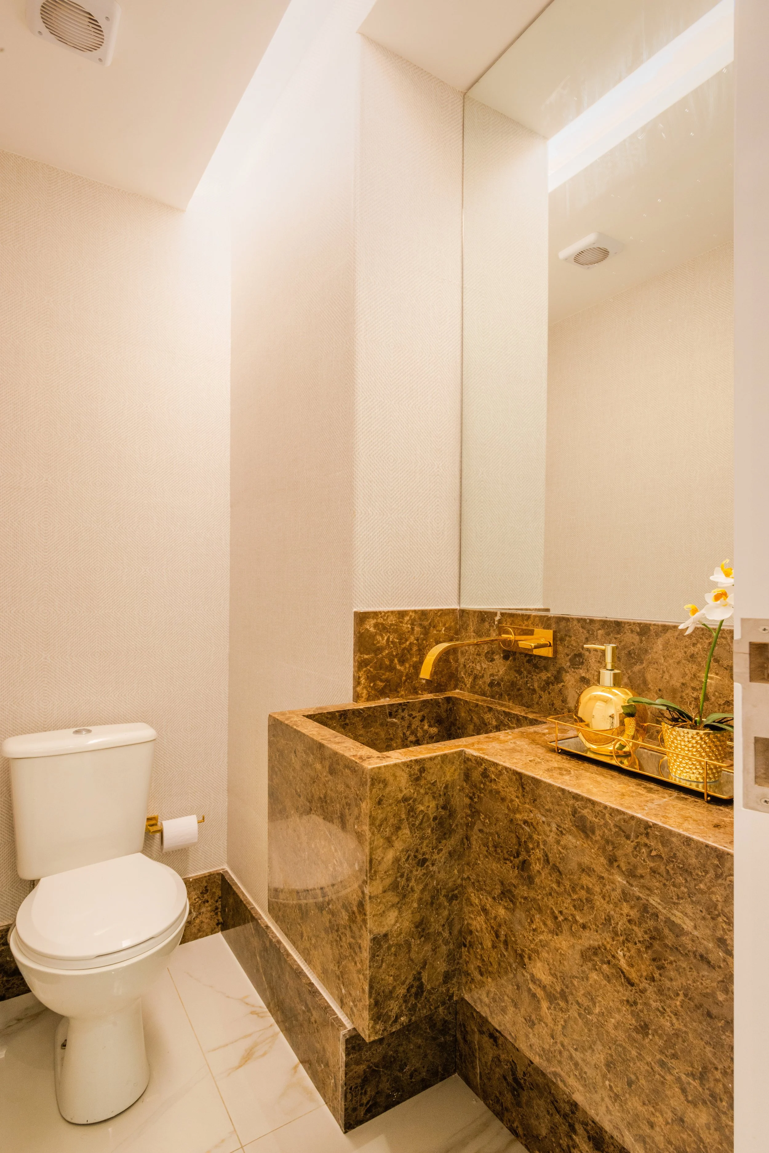 Modern bathroom with beige walls, a white toilet, and a brown marble sink area with gold fixtures and decoration, including a potted orchid.
