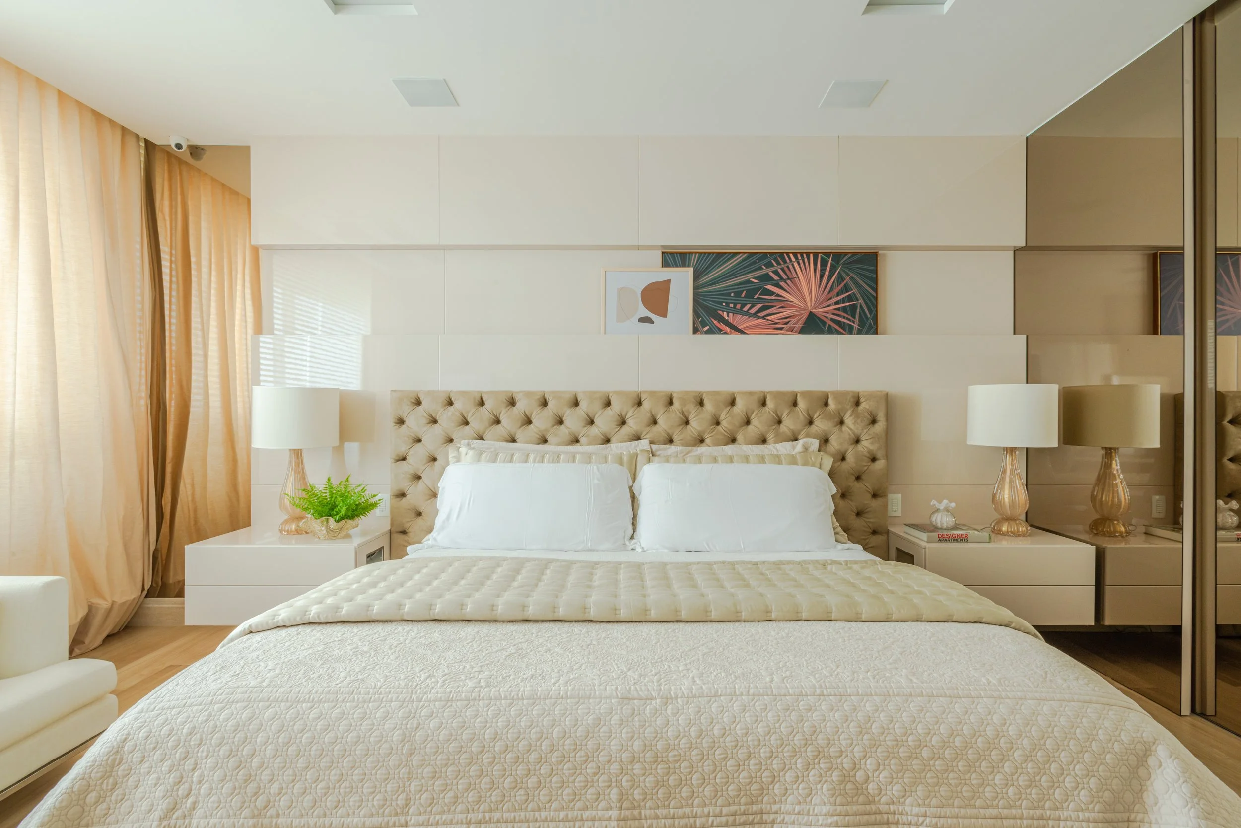 A bedroom with a large bed featuring a tufted beige headboard, white bedding, and cream-colored pillows. There are two bedside tables with matching lamps, one on each side of the bed. The left side has a potted green plant, and the right side has a s