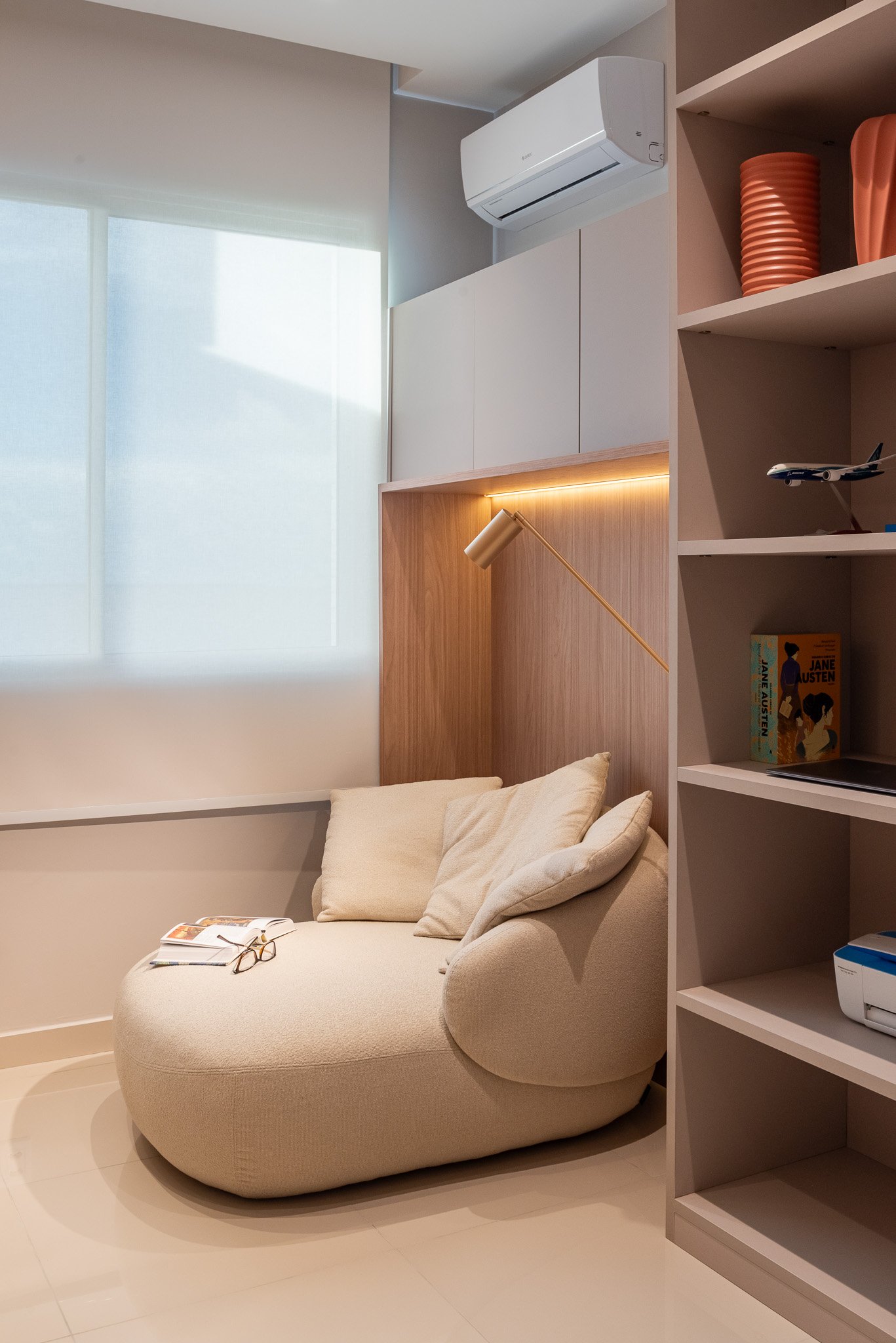 A cozy beige armchair with pillows, placed near a window with sheer curtains, in a modern living room with a wooden headboard and shelving with decorative items.