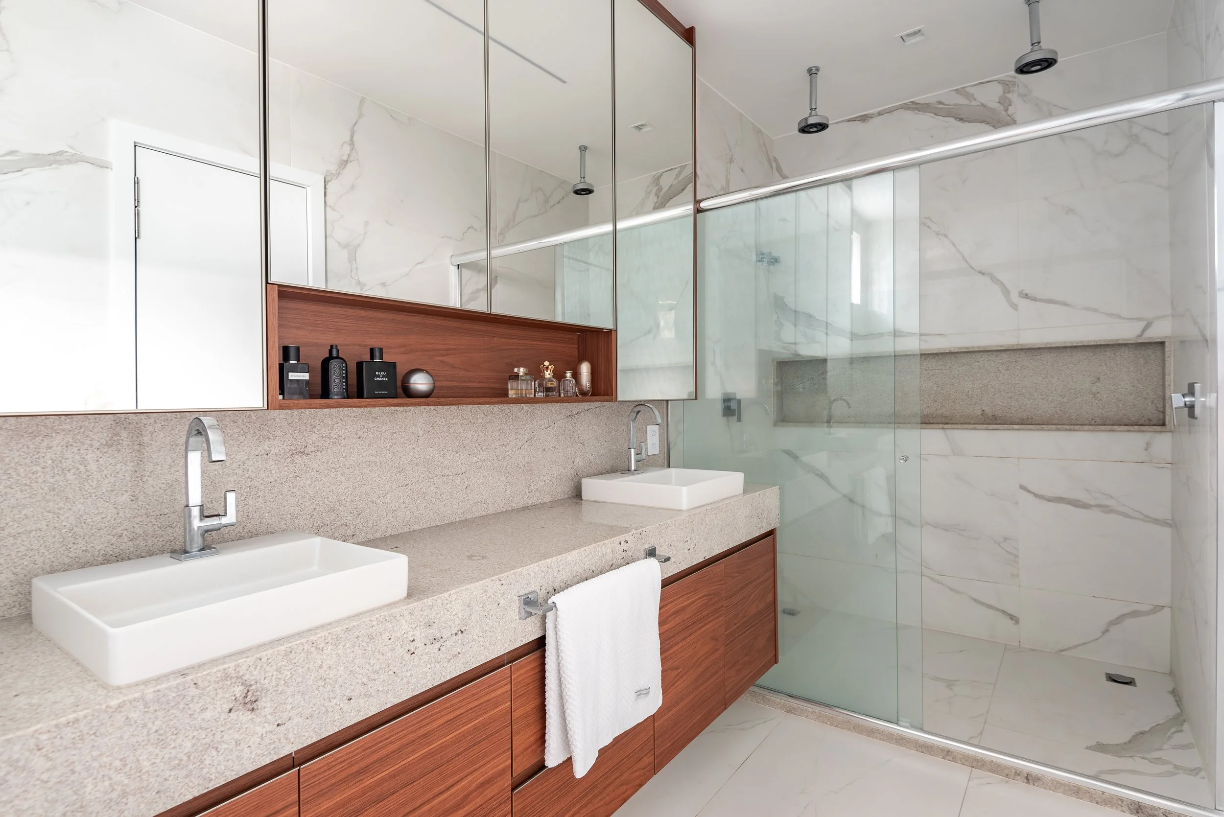 Modern bathroom with double vessel sinks, granite countertop, wooden cabinetry, large mirror, marble walls, and glass shower with built-in shelf.