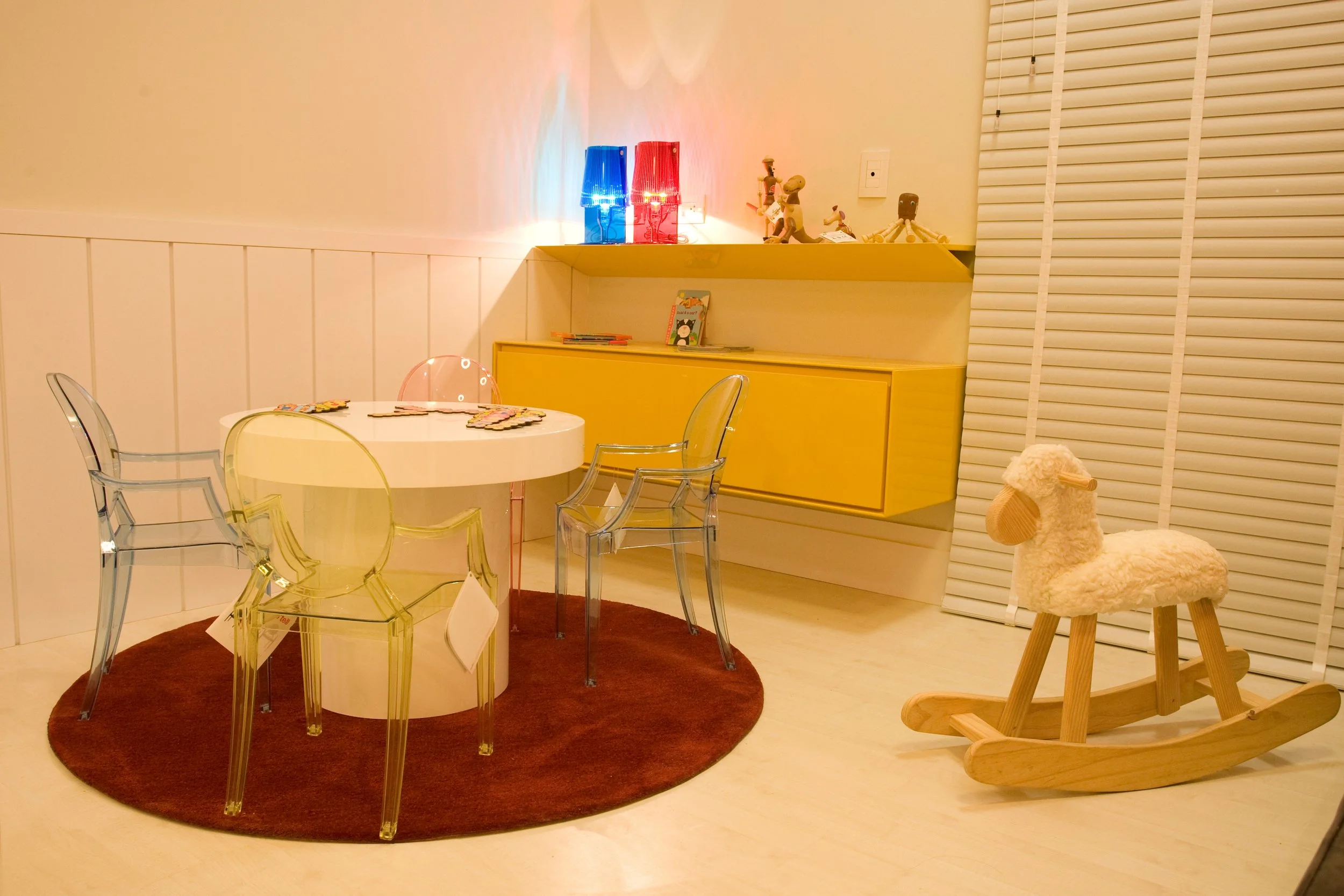 Kids' playroom with a white round table surrounded by transparent and yellow chairs on a red round rug, a yellow wall-mounted cabinet, a wooden rocking horse, and toys on the shelf, illuminated with warm lighting.