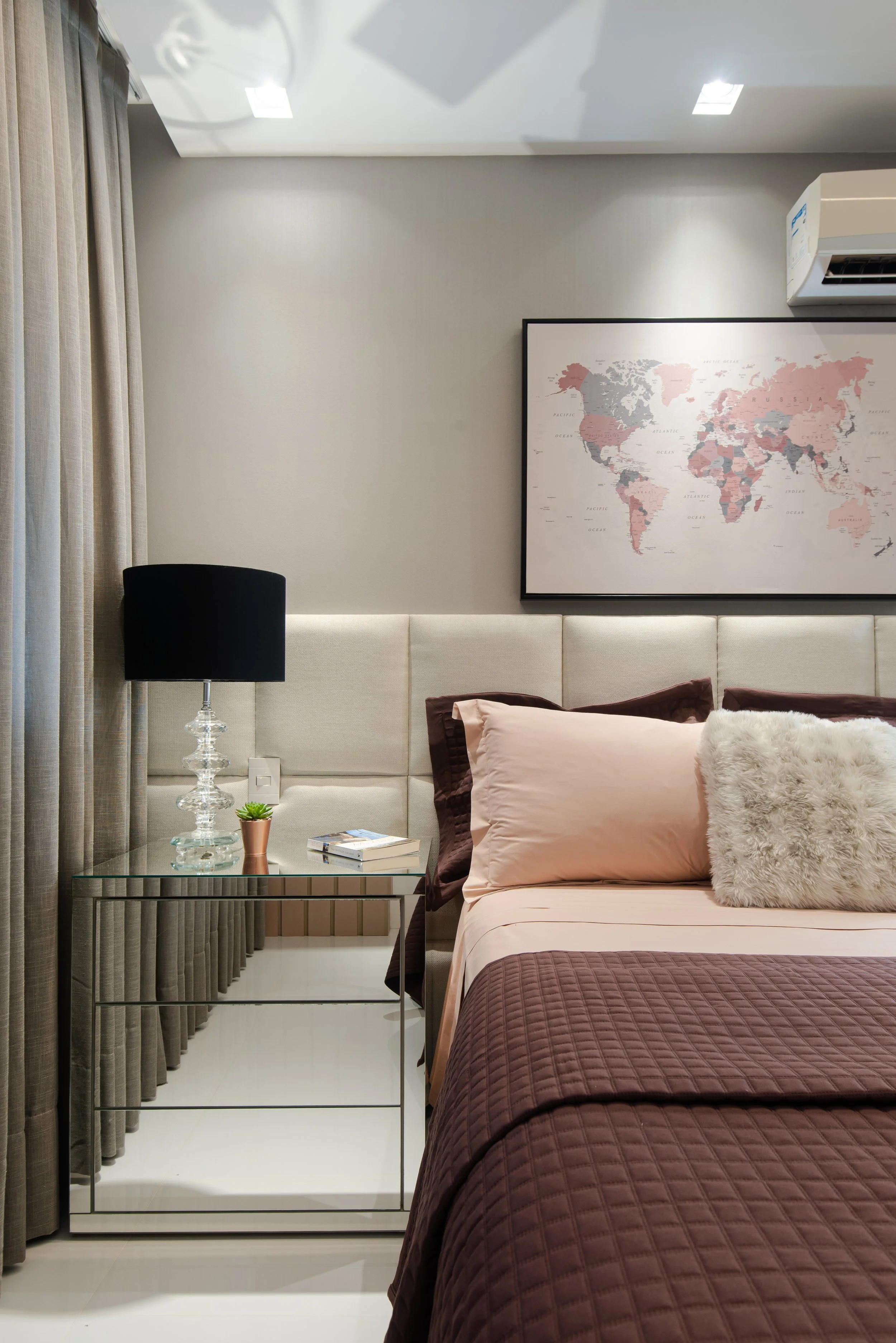 Modern bedroom with beige upholstered headboard, pink and brown bedding, a mirrored nightstand with a black lamp, a small plant, books, and a world map poster on the wall.