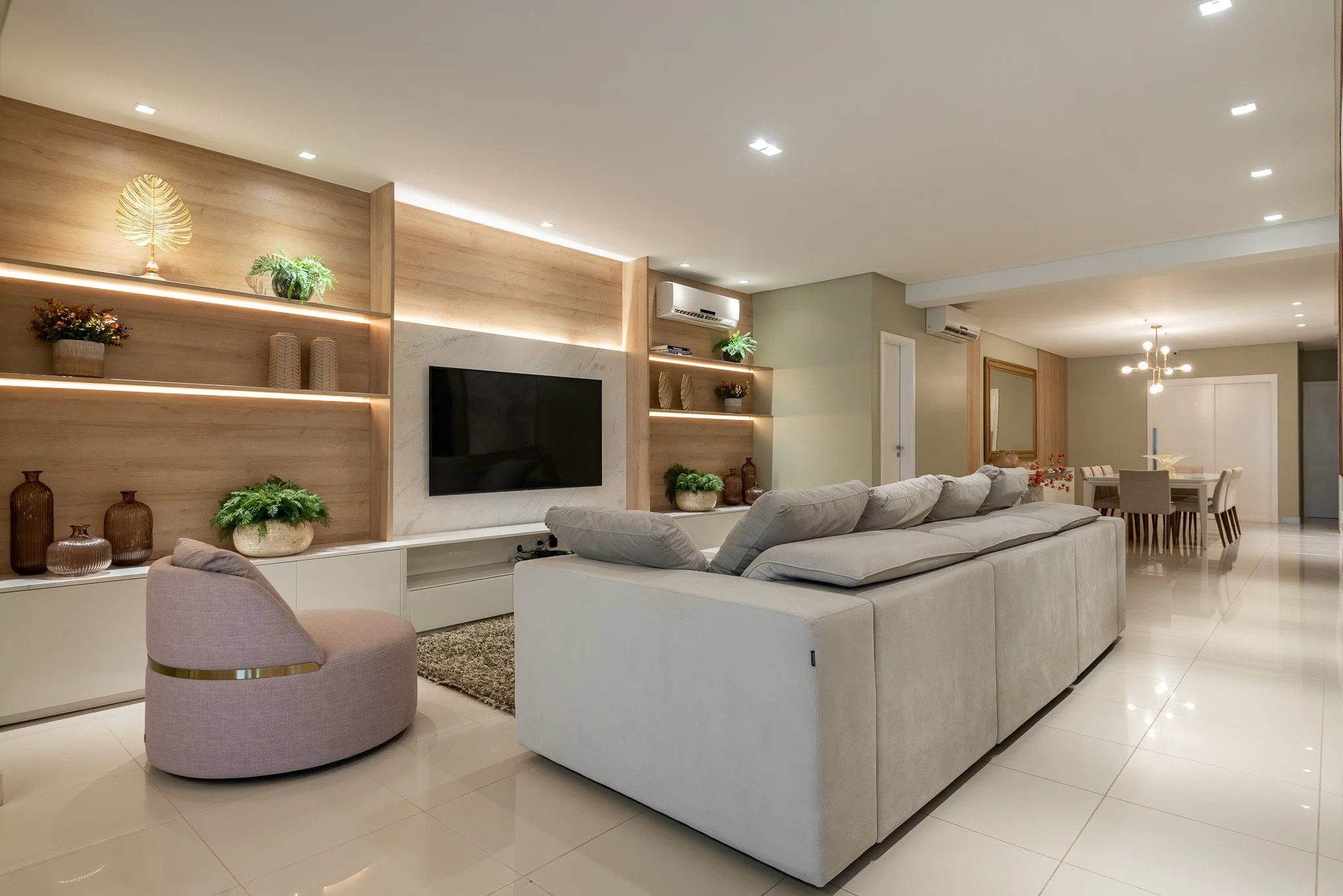 Modern living room with white sofa, circular pink armchair, and decorative shelves with plants and vases, open to dining area with chandelier.