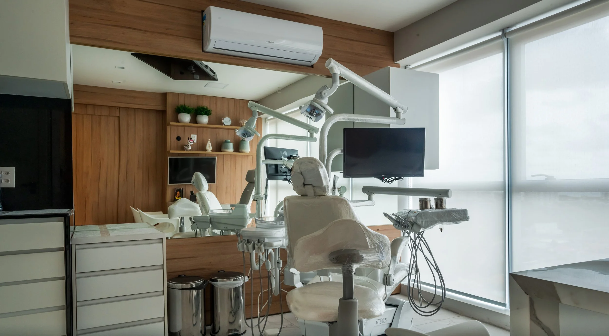 Dental clinic treatment room with dental chair, overhead lights, monitors, and cabinetry, adjacent to a large window with blinds.