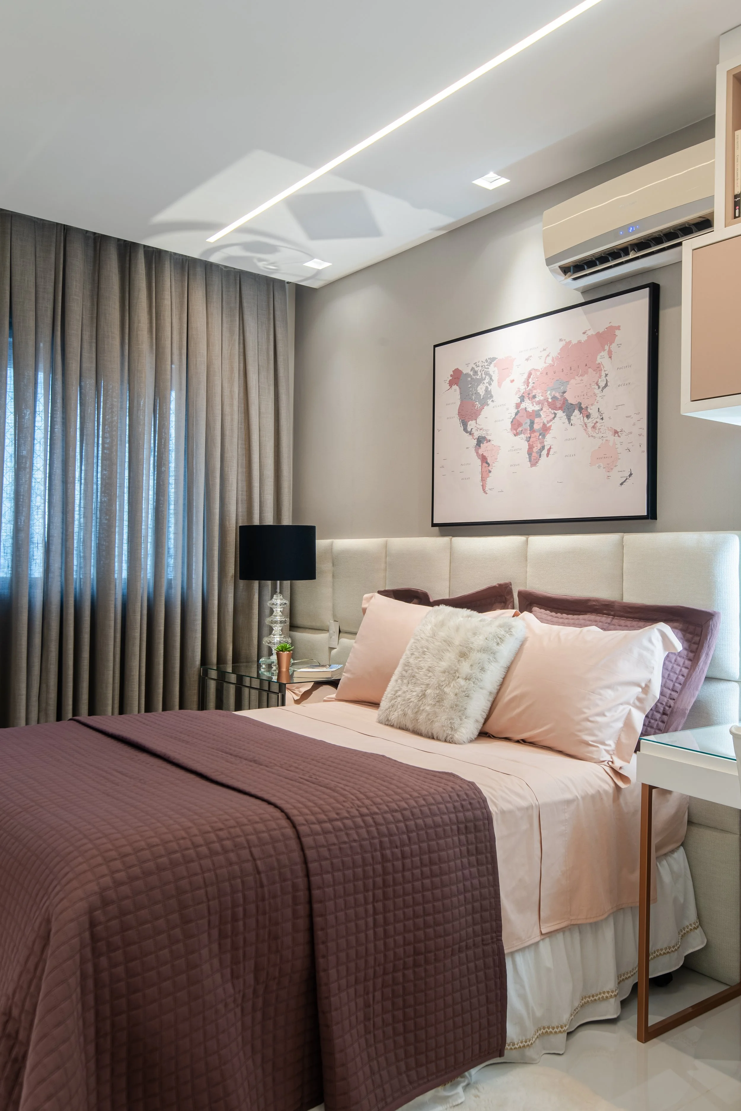 A bedroom with a large bed dressed in pink and brown bedding, a black bedside lamp on a glass side table, a world map in pinks and grays on the wall, beige curtains covering a window, and an air conditioning unit above the map.