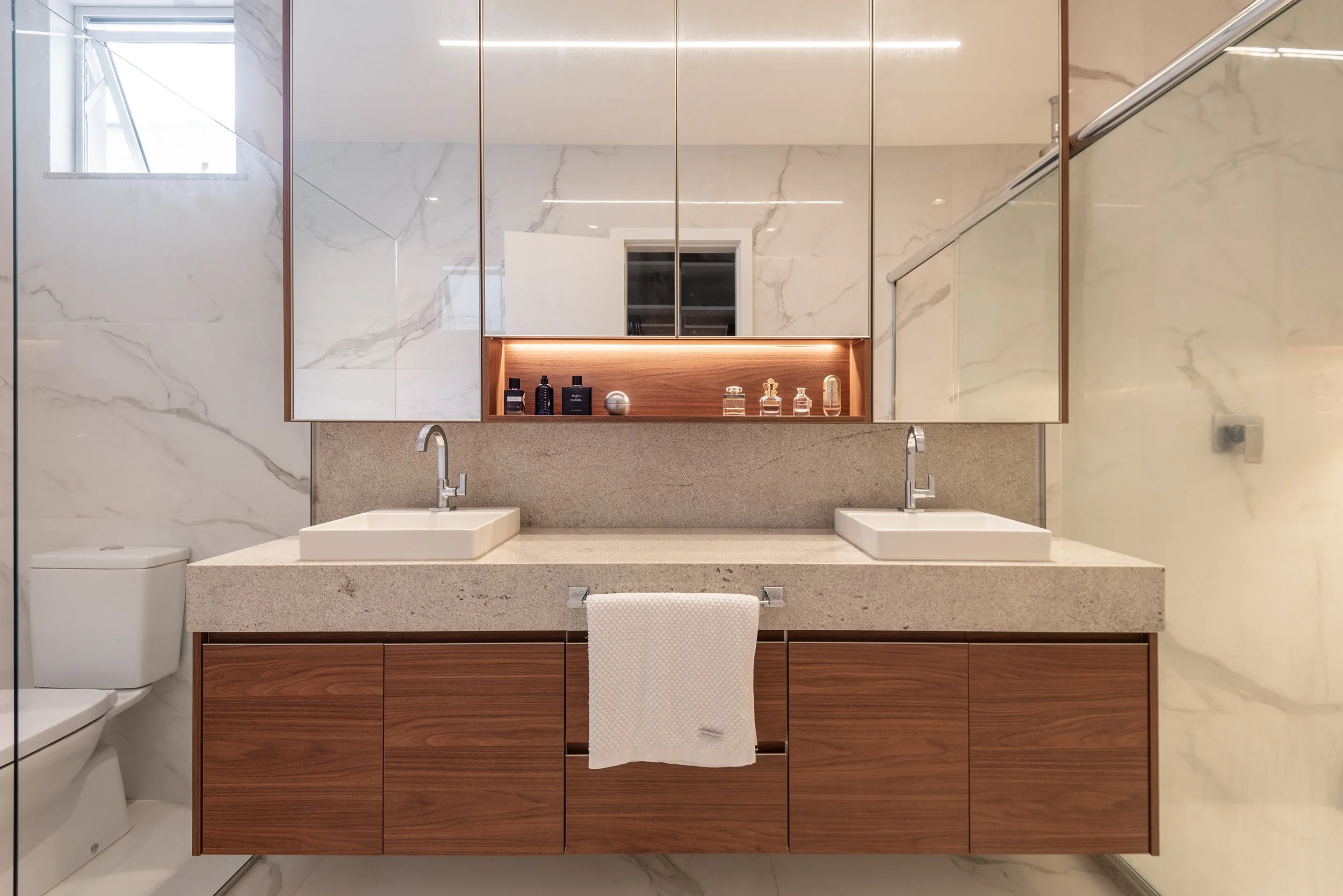 Modern bathroom with double vanity, two rectangular vessel sinks, wood cabinetry, large mirror, marble walls, and a towel hanging on the cabinet handle.