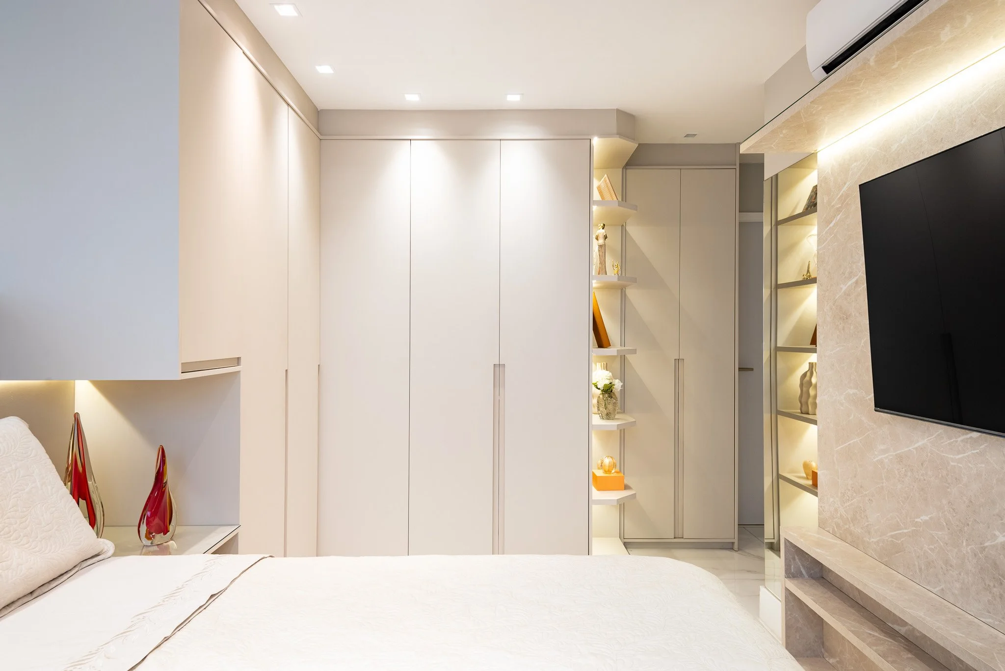 Modern bedroom with white built-in wardrobes, decorative shelves with lights, a TV mounted on a beige marble wall, and a bed with cream-colored bedding.