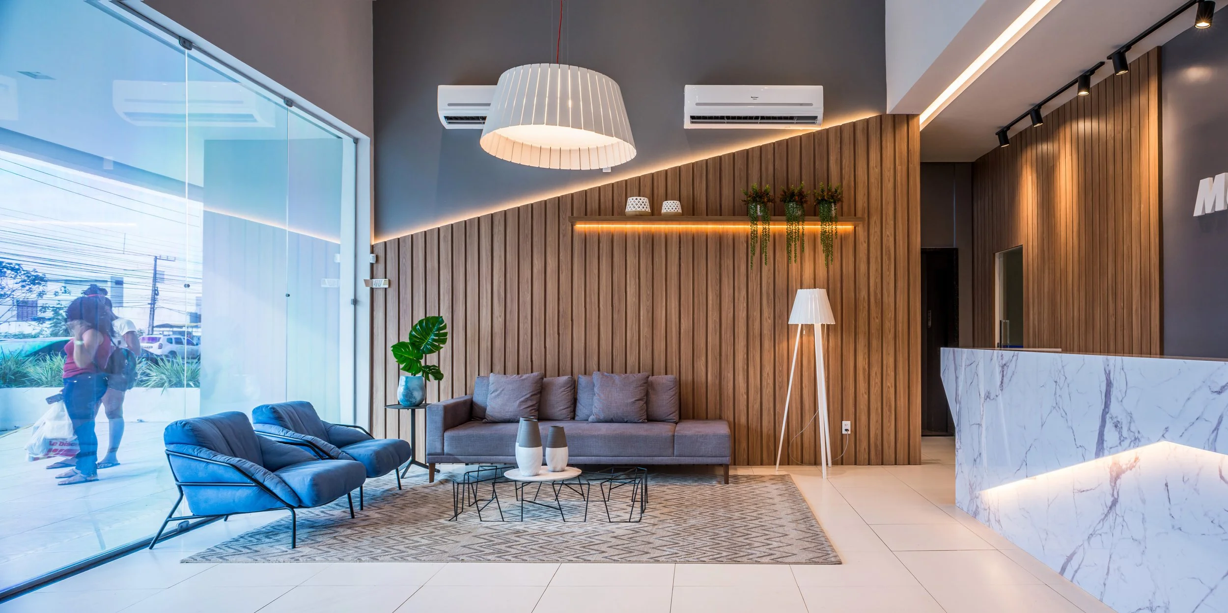 Modern hotel lobby with wooden accent wall, gray sofa, black chairs, a patterned rug, potted plant, white floor lamp, and marble reception desk, with large glass windows and ceiling lighting.