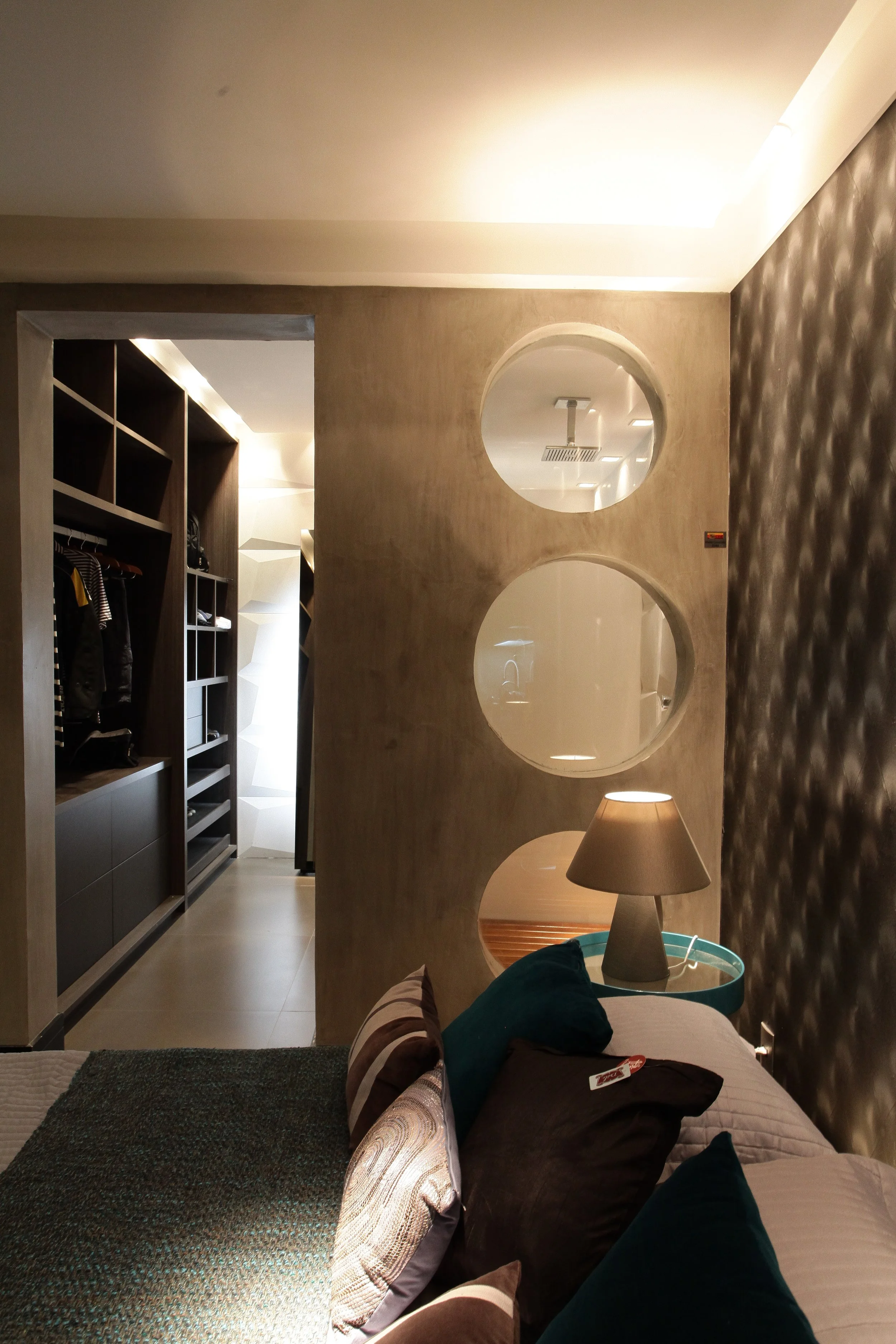 A bedroom with a bed, pillows, a bedside lamp, a decorative wall, and a circular mirror on the wall with circles cut out, revealing a closet and a ceiling with lights and a shower head.