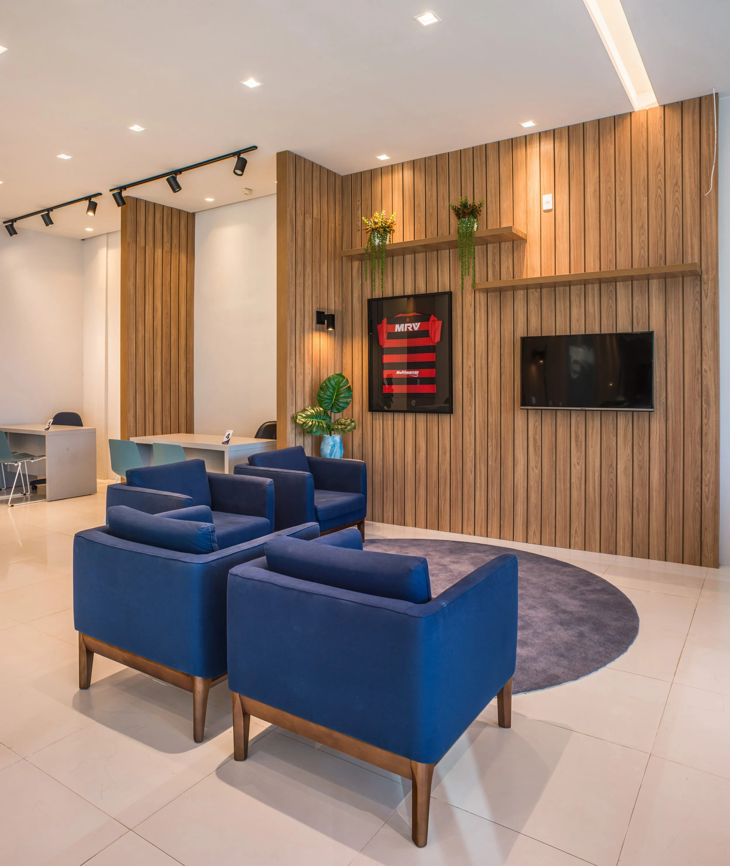 A modern lobby area with blue armchairs, a wooden wall with shelves, potted plants, a mounted TV, and a framed sports jersey.