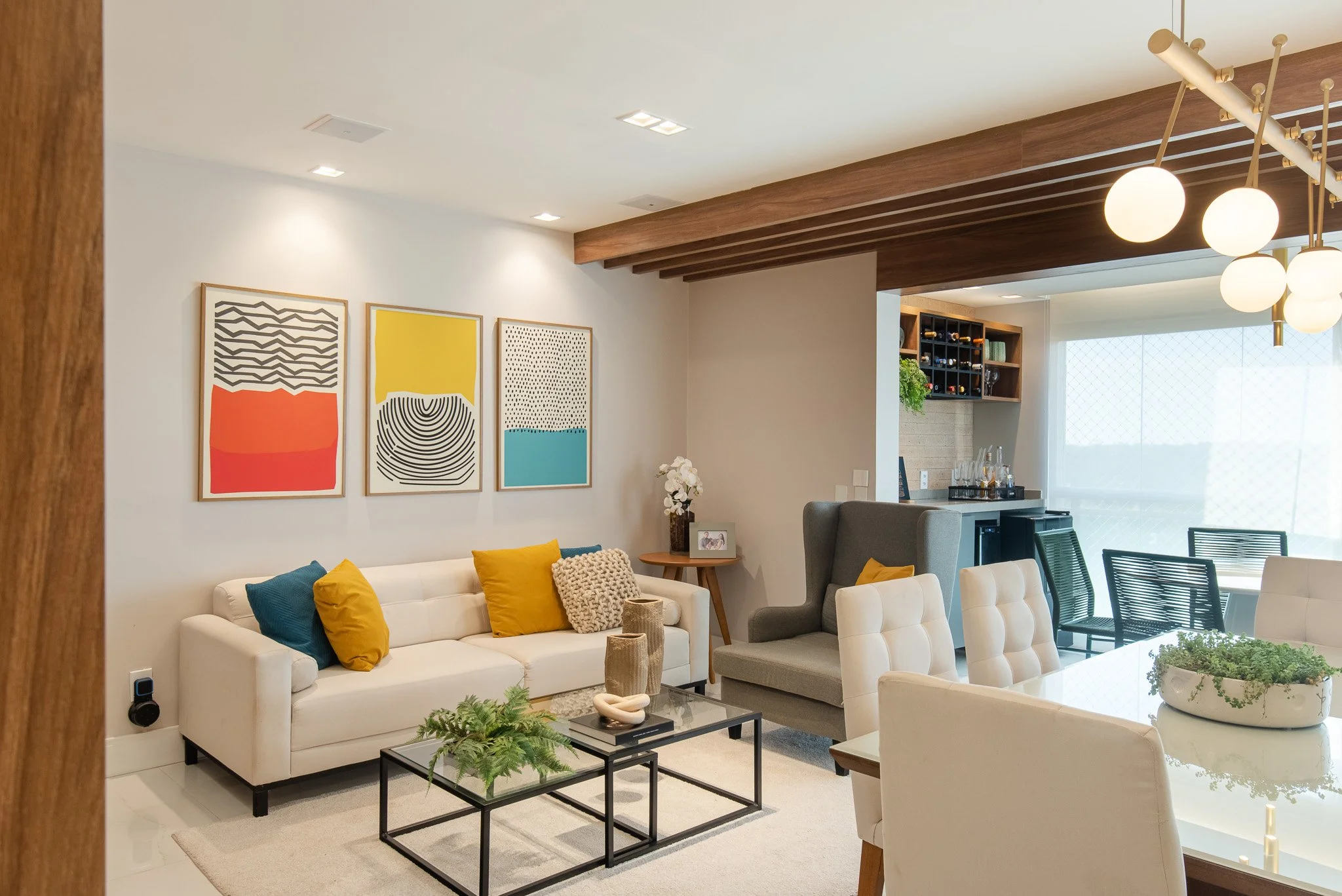 Modern living room with white sofa, colorful cushions, abstract artwork on the wall, glass coffee table, and adjacent dining area, with a bar area and large window with sheer curtains.