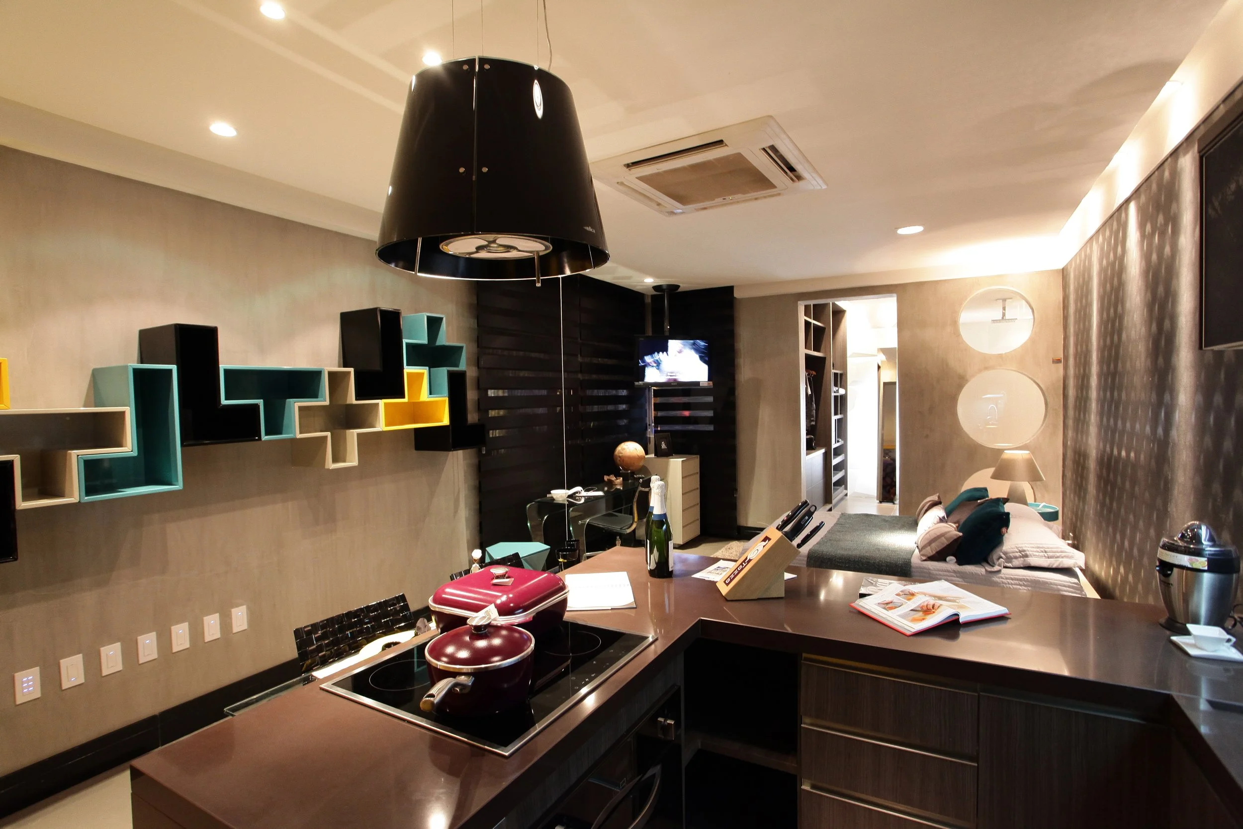 A modern kitchen and living room with dark wood and colorful geometric wall shelves, a large black hanging lamp, a counter with pots, a book, and a champagne bottle, a small TV, a bed with pillows, and a patterned wall with circular mirrors.