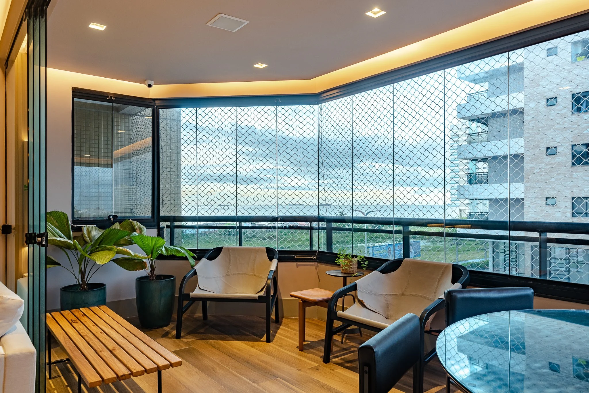 Indoor balcony area with two white armchairs, black frames, small side table with a plant, potted plants, a wooden bench, a glass table, and large windows with a safety net, overlooking city buildings and a cloudy sky.