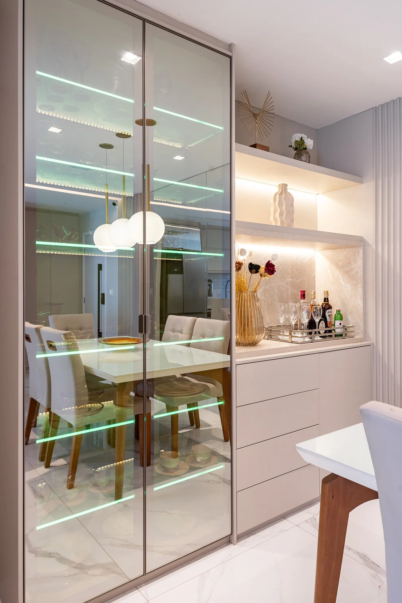 Modern dining area with a glass display cabinet reflecting a dining table and chairs, decorative shelves with vases and art pieces, and a marble countertop with a tray of liquor bottles and glasses.