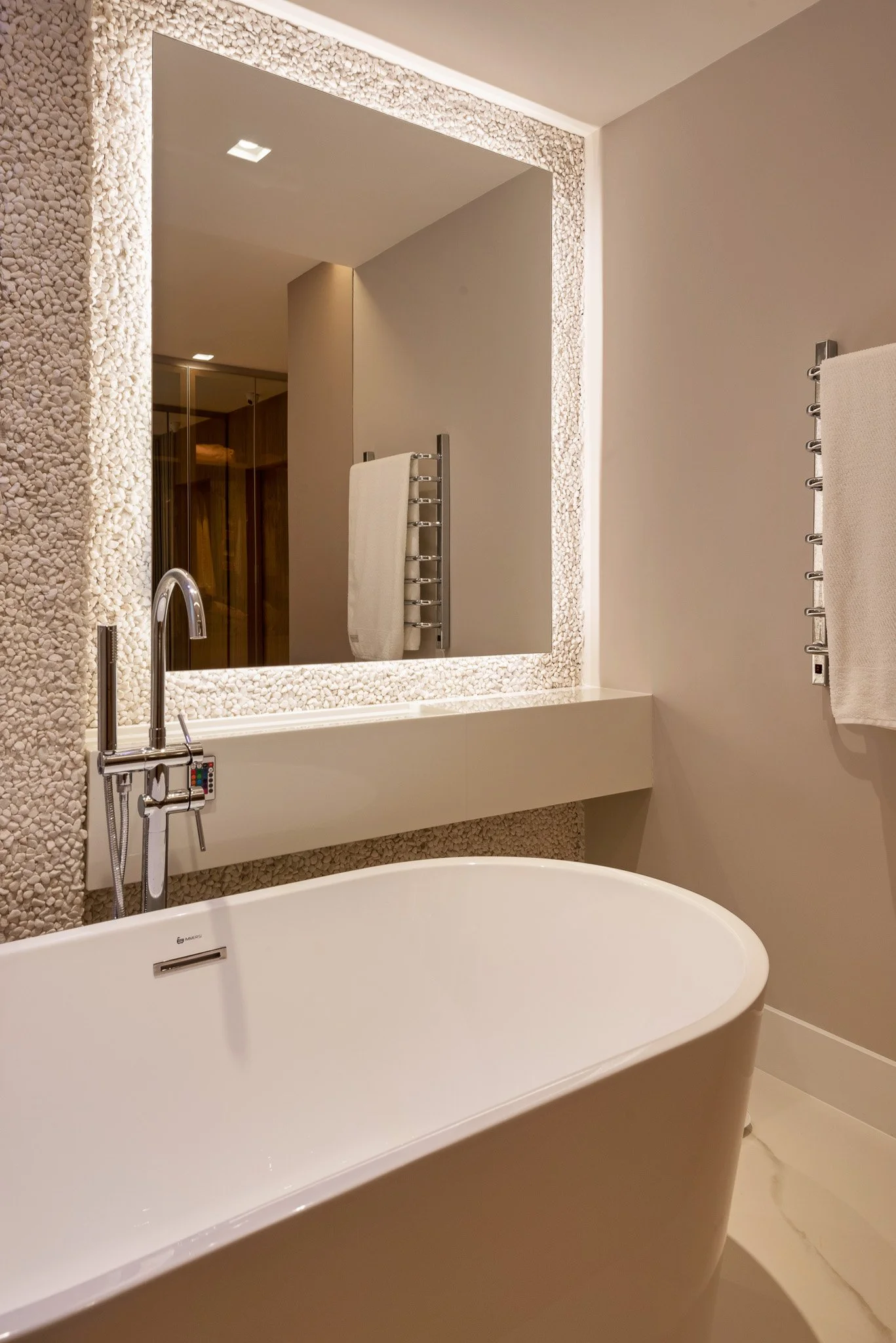 Modern bathroom with a white bathtub, large mirror with a pebble texture border, beige walls, a chrome heated towel rail, and a beige towel hanging on the side.