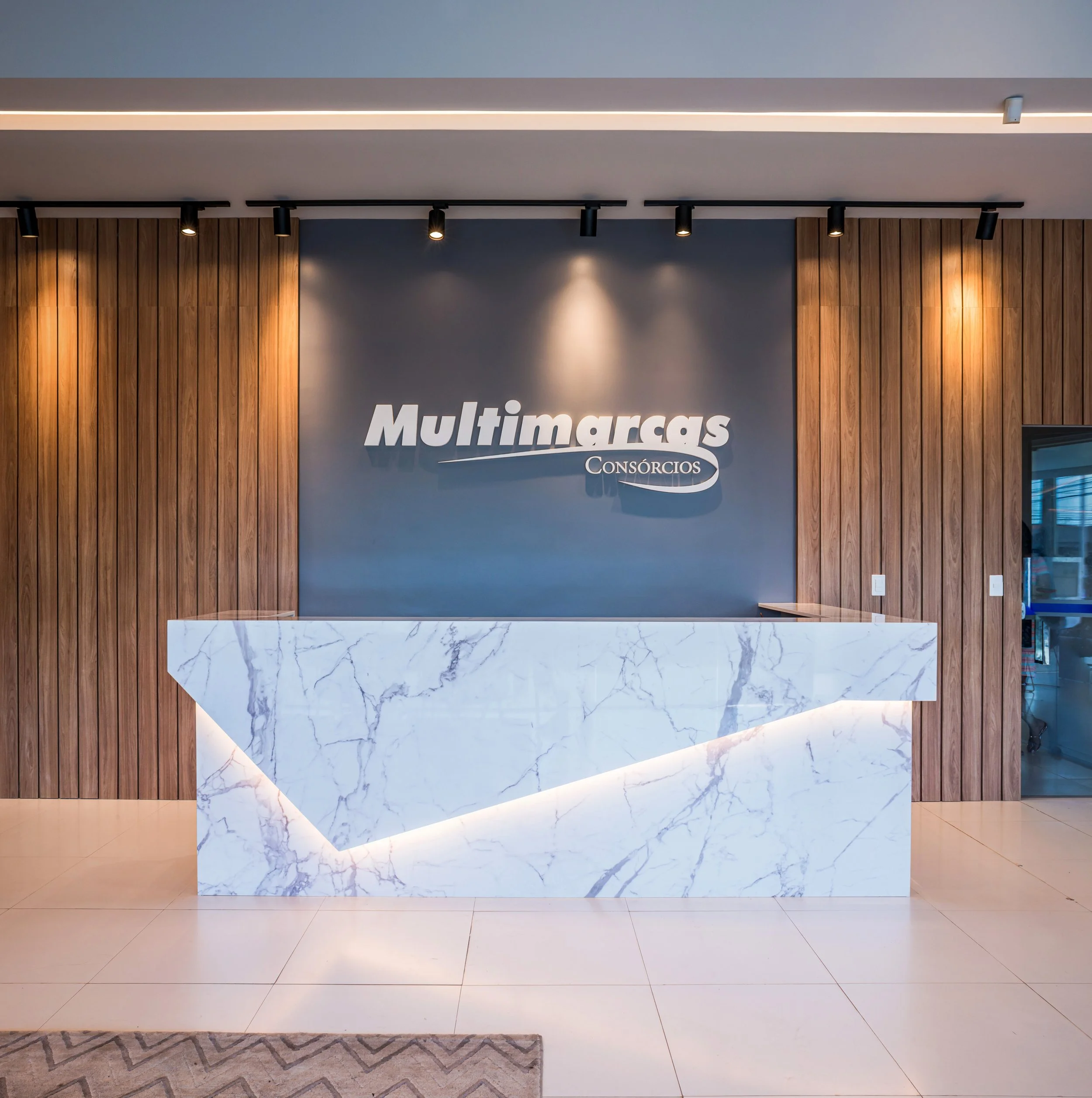 Reception desk made of white marble with gray veins, in front of a dark gray wall with the company's name 'Multimarcas Consórcios' in silver letters, flanked by wooden wall panels, with ceiling lights above.