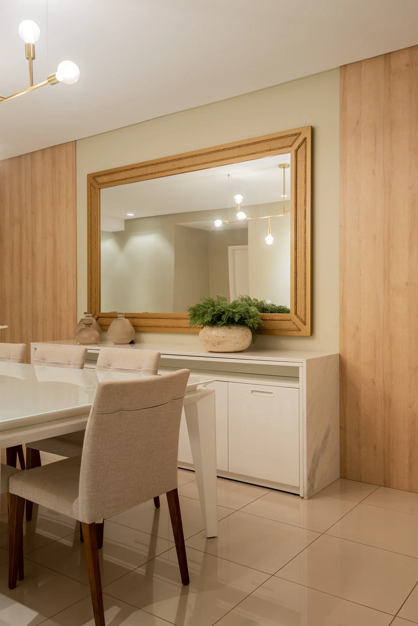 A dining room with a large mirror, beige upholstered chairs, a white table, a white cabinet, a potted plant, and a modern light fixture.