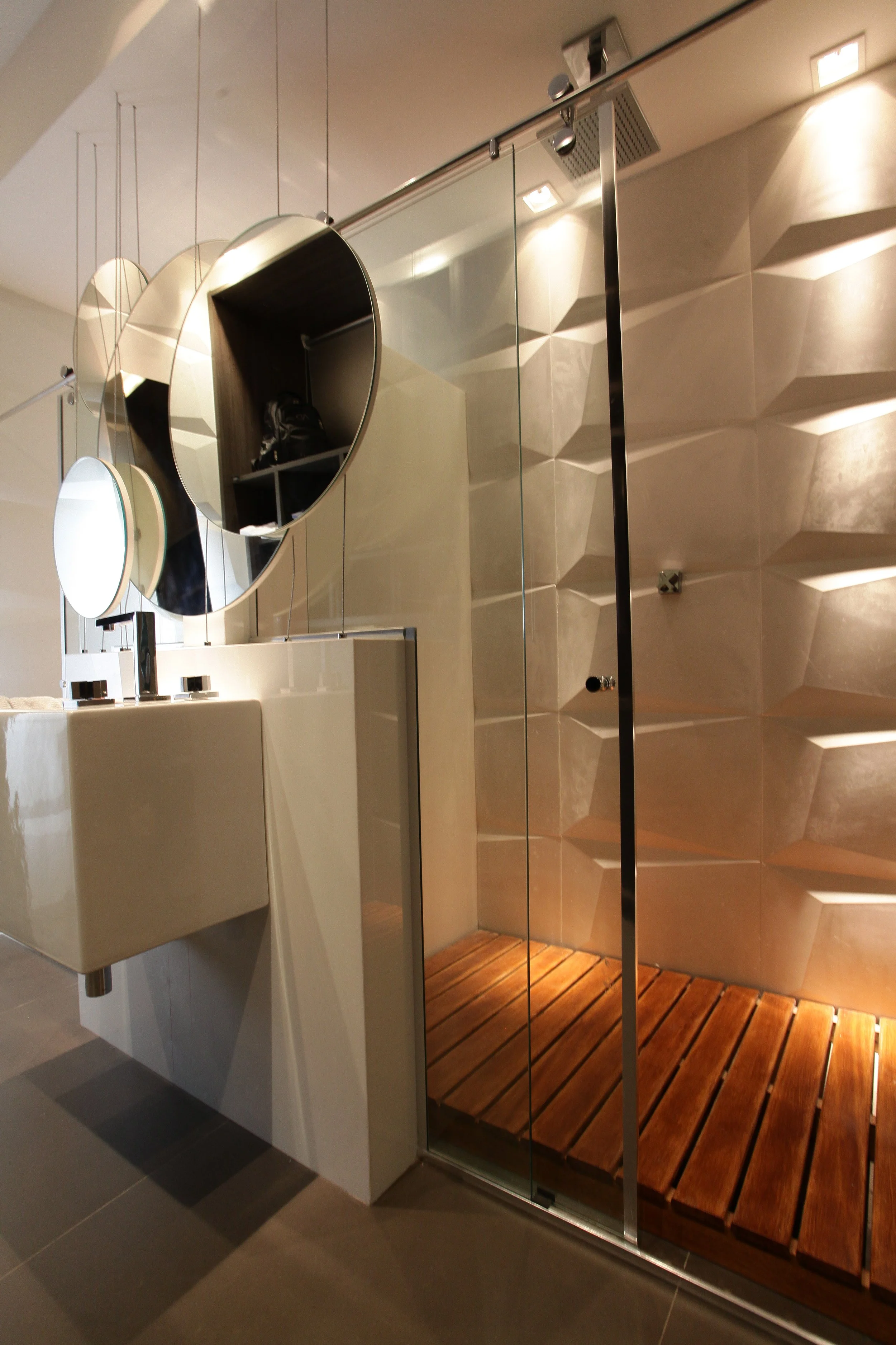 Modern bathroom with a glass shower enclosure featuring a wooden shower platform and a wall with a textured geometric pattern. A white sink with a faucet is seen in front of a set of circular mirrors.