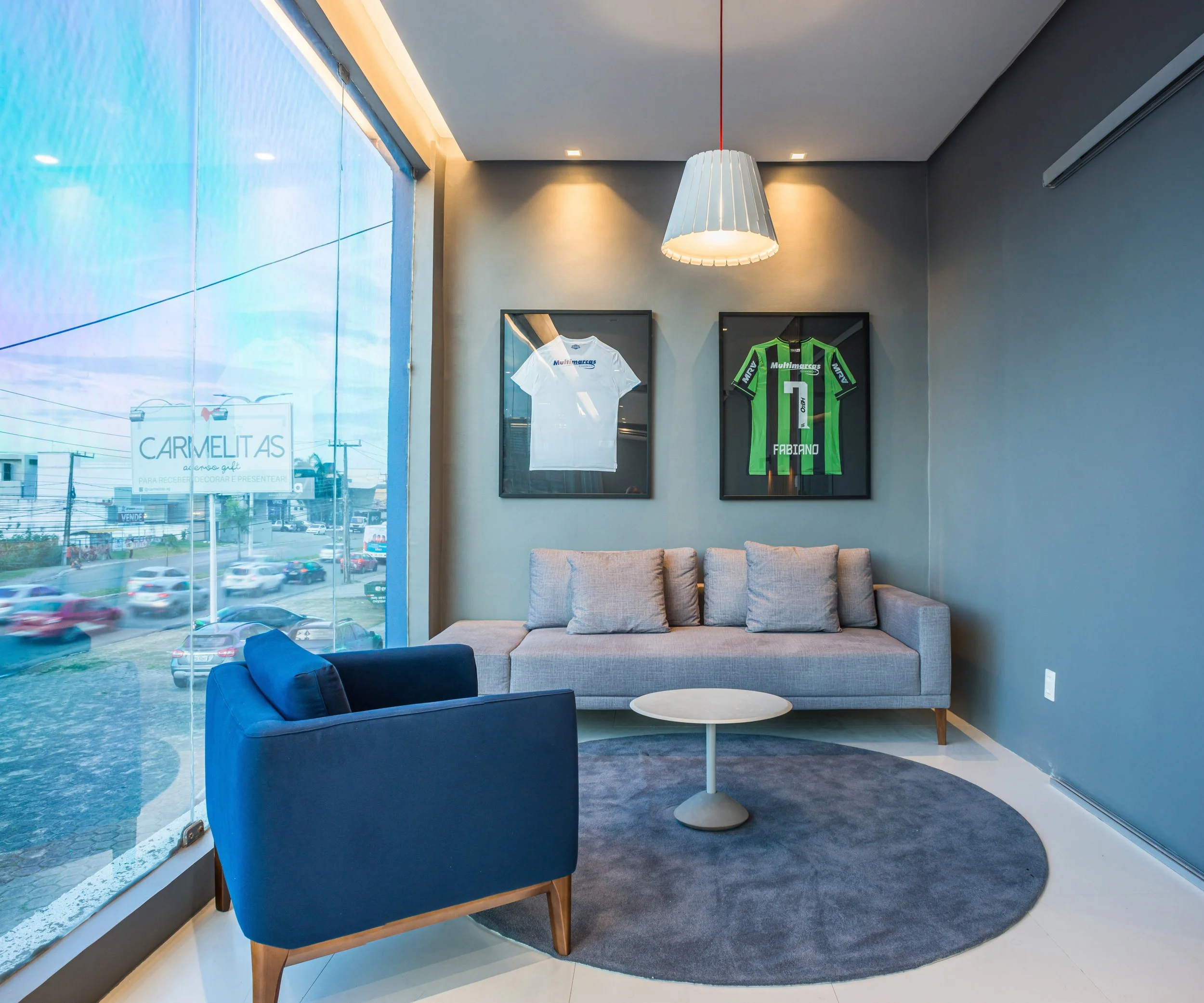 Modern interior waiting area with a large window, a gray and blue sofa, a navy armchair, a white round coffee table, framed soccer jerseys on the wall, and a hanging white lamp.