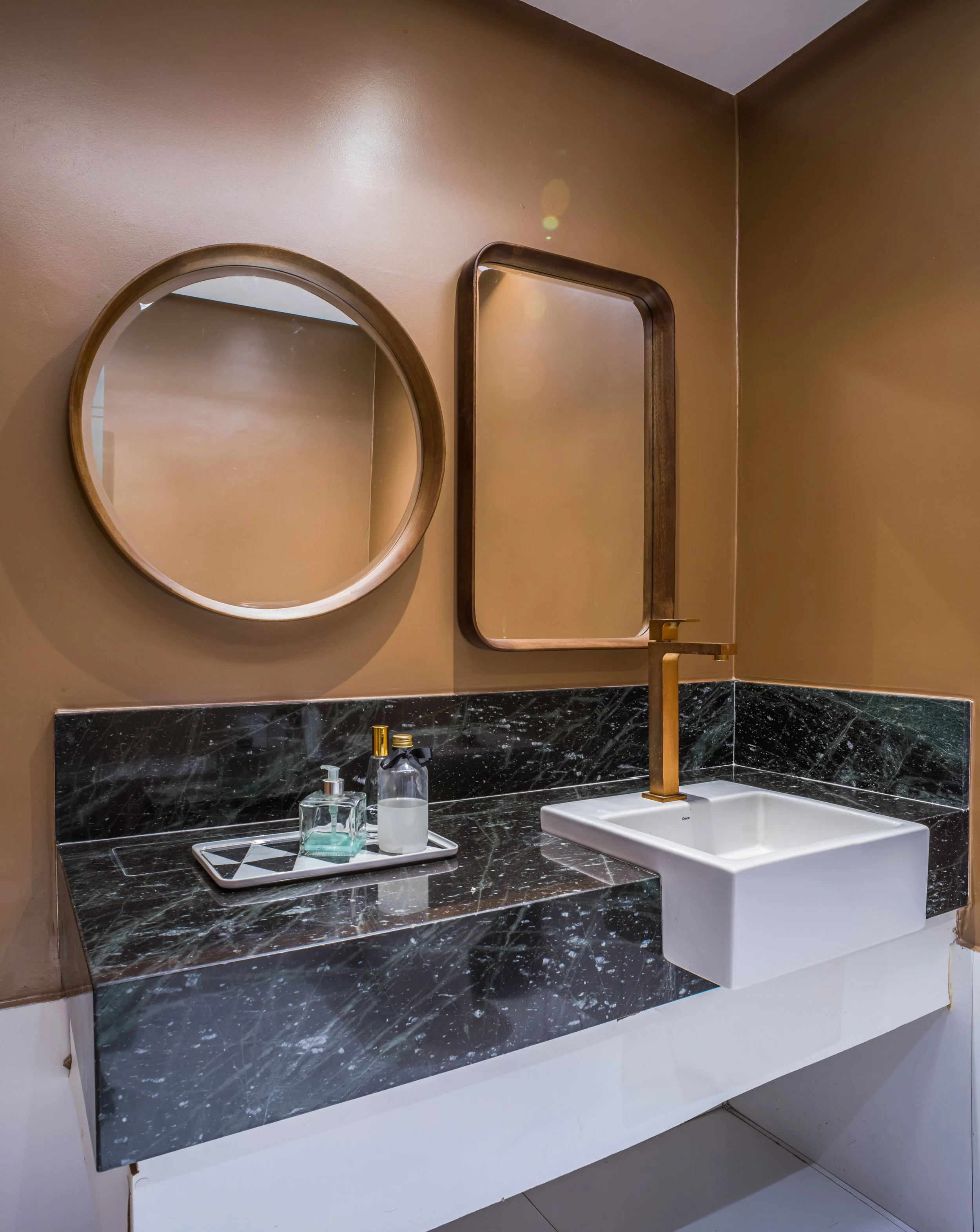Modern bathroom with a black marble countertop, rectangular white sink, gold faucet, round and rectangular mirrors on a beige wall, and bottles of soap on a tray.