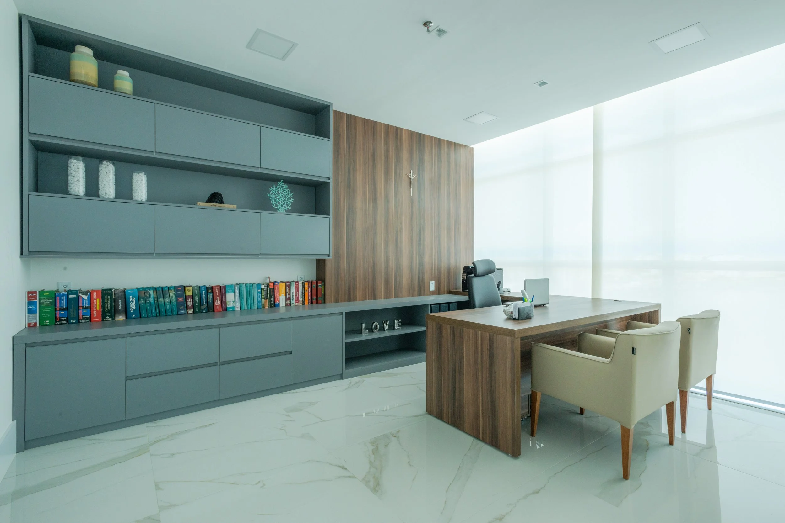 Modern office with grey bookshelf, wooden desk, beige chairs, and large window with blinds