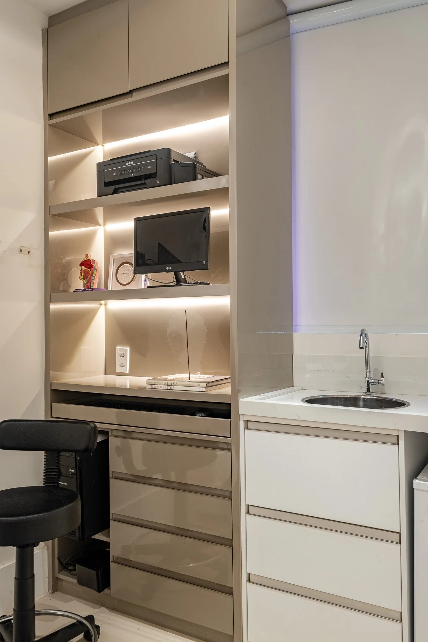 View of a kitchenette with a small sink on the right, beige cabinets, and open shelves with a printer, a small TV, a framed photo, decorative figurines, and books, with a black office chair in the foreground.